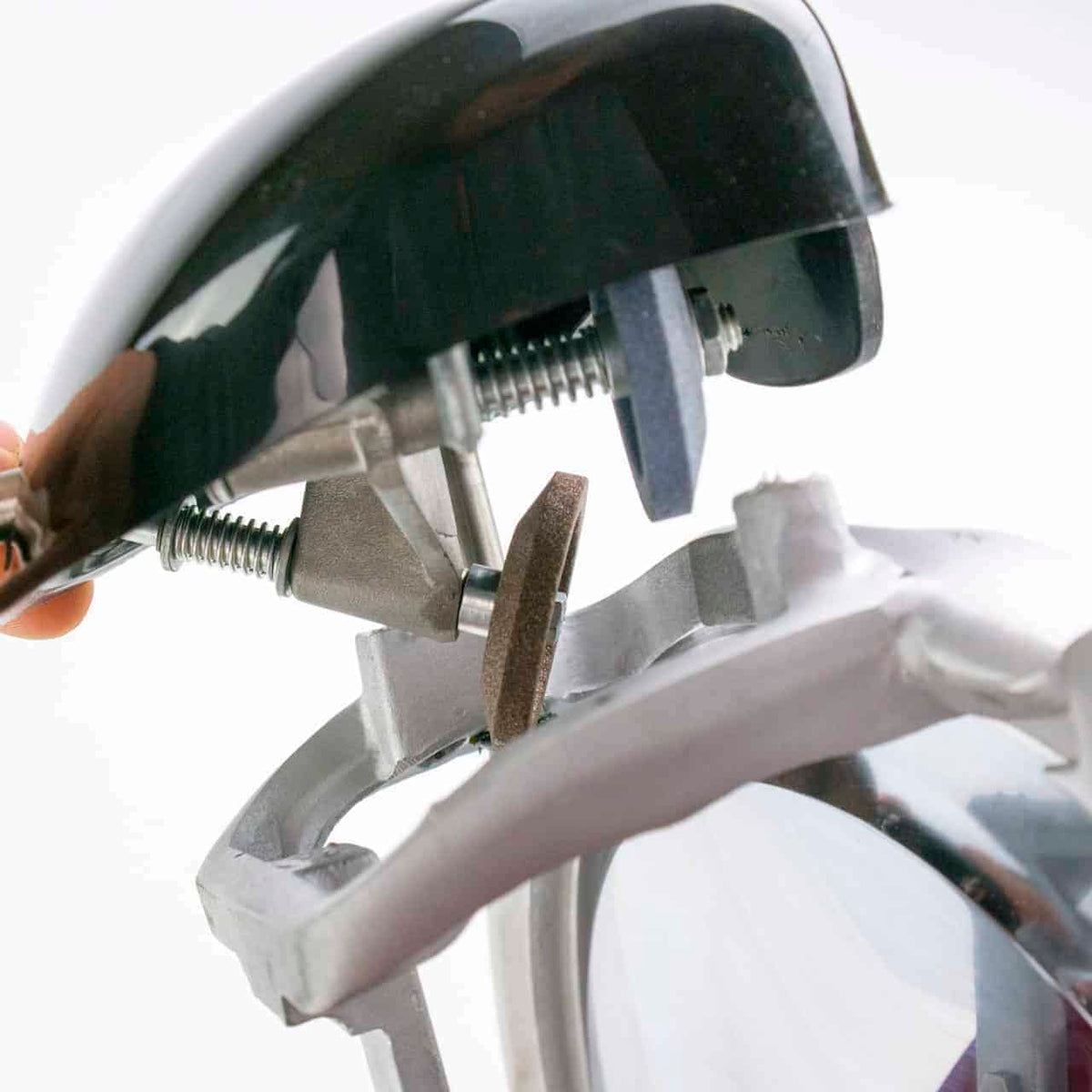Nahaufnahme einer Maschinenkomponente mit einem durch Schrauben befestigten Klemmmechanismus und einer Schleifscheibe. Der Aufbau zeigt, wie Metallteile eingestellt werden, wahrscheinlich für präzise mechanische Arbeiten oder die Montage. Eine Hand ist teilweise sichtbar, die den oberen Teil hält.