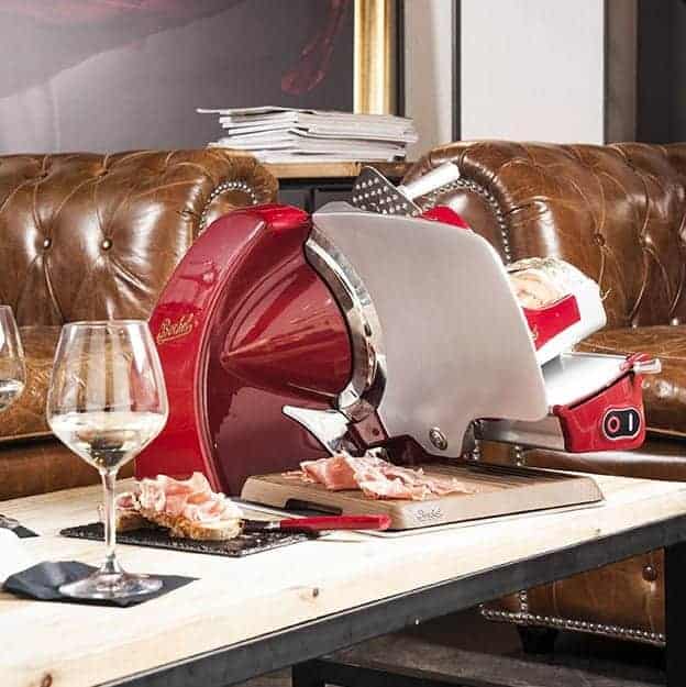 Eine rot-silberne Aufschnittmaschine steht auf einem hölzernen Couchtisch vor einem braunen Ledersofa. Die Maschine schneidet Schinkenscheiben, und auf dem Tisch stehen zwei Weingläser, die teilweise mit Weißwein gefüllt sind, sowie einige Schinkenscheiben auf einer schwarzen Schieferplatte.