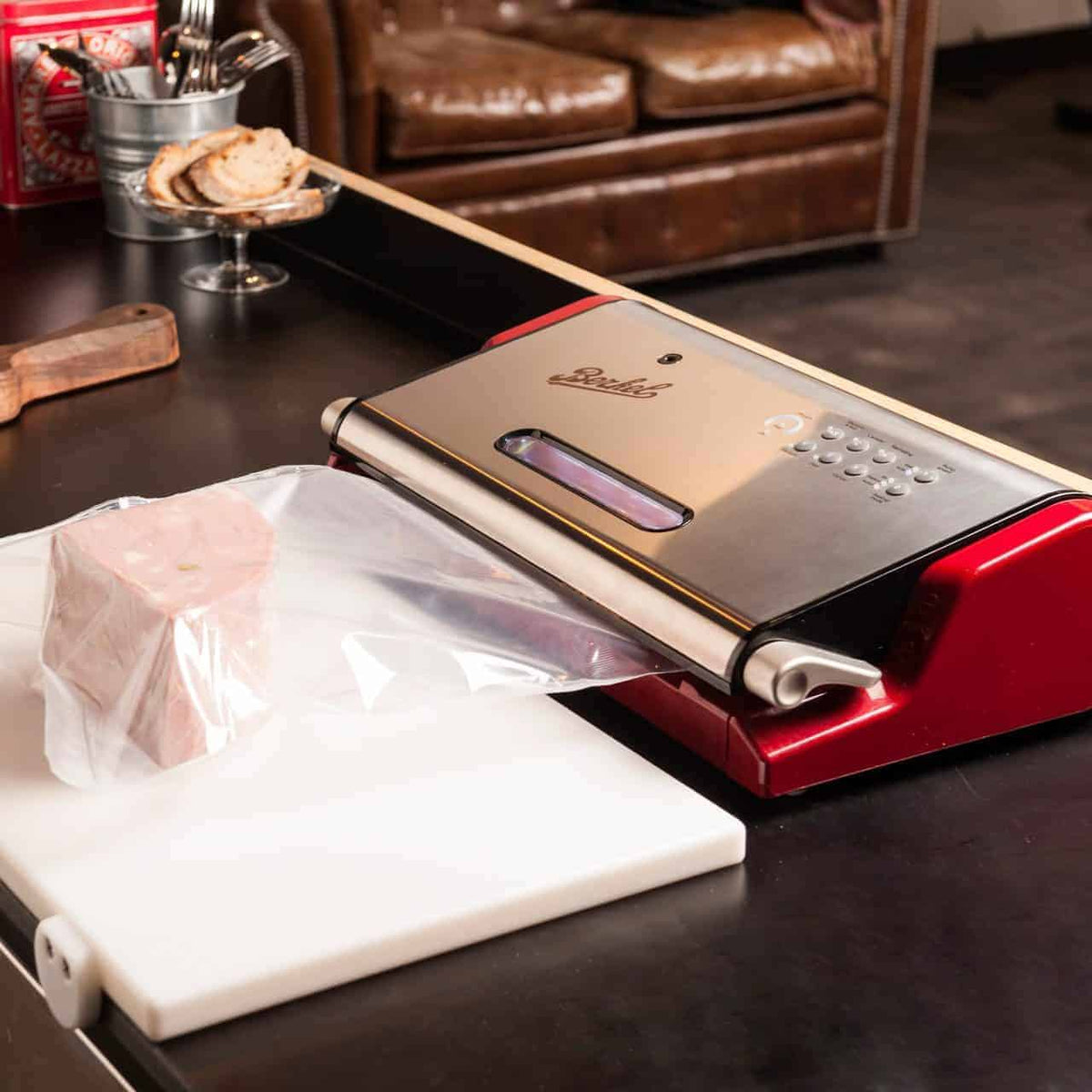 Eine Berkel Vacuum Vakuummaschine steht auf einer Küchentheke und versiegelt ein Stück Fleisch in einer Plastiktüte. Im Hintergrund sind ein Schneidebrett mit geschnittenem Brot und ein Messer zu sehen, zusammen mit einem braunen Ledersofa und anderen Küchenutensilien.