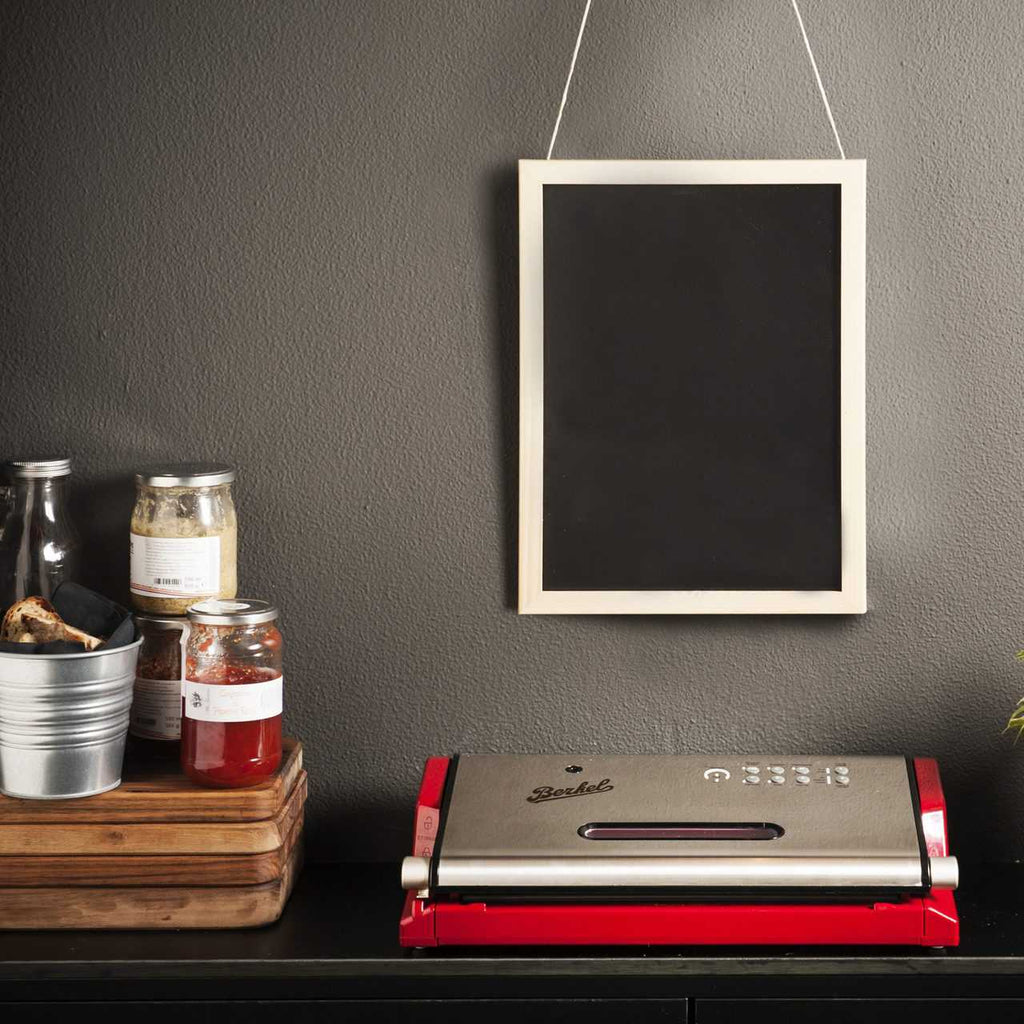 Eine Küchenszene mit einem Berkel Vacuum, Vakuummaschine in Rot und Silber auf einer schwarzen Arbeitsplatte. Darüber hängt eine kleine Tafel mit weißem Rahmen an einer dunkelgrauen Wand. Links davon stehen auf einem Holzpodest Gläser und ein Metallbehälter mit Küchenutensilien.