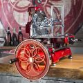 Eine Vintage-Aufschnittmaschine Berkel Volano B114 Rot mit passgenauem Einlegebrett, ausgestattet mit einer Handkurbel und aufwendigen Mustern, steht auf einer Holztheke. Im Hintergrund sind einige Flaschen und eine große runde Wanddekoration zu sehen, die zu einer rustikalen Atmosphäre beitragen.