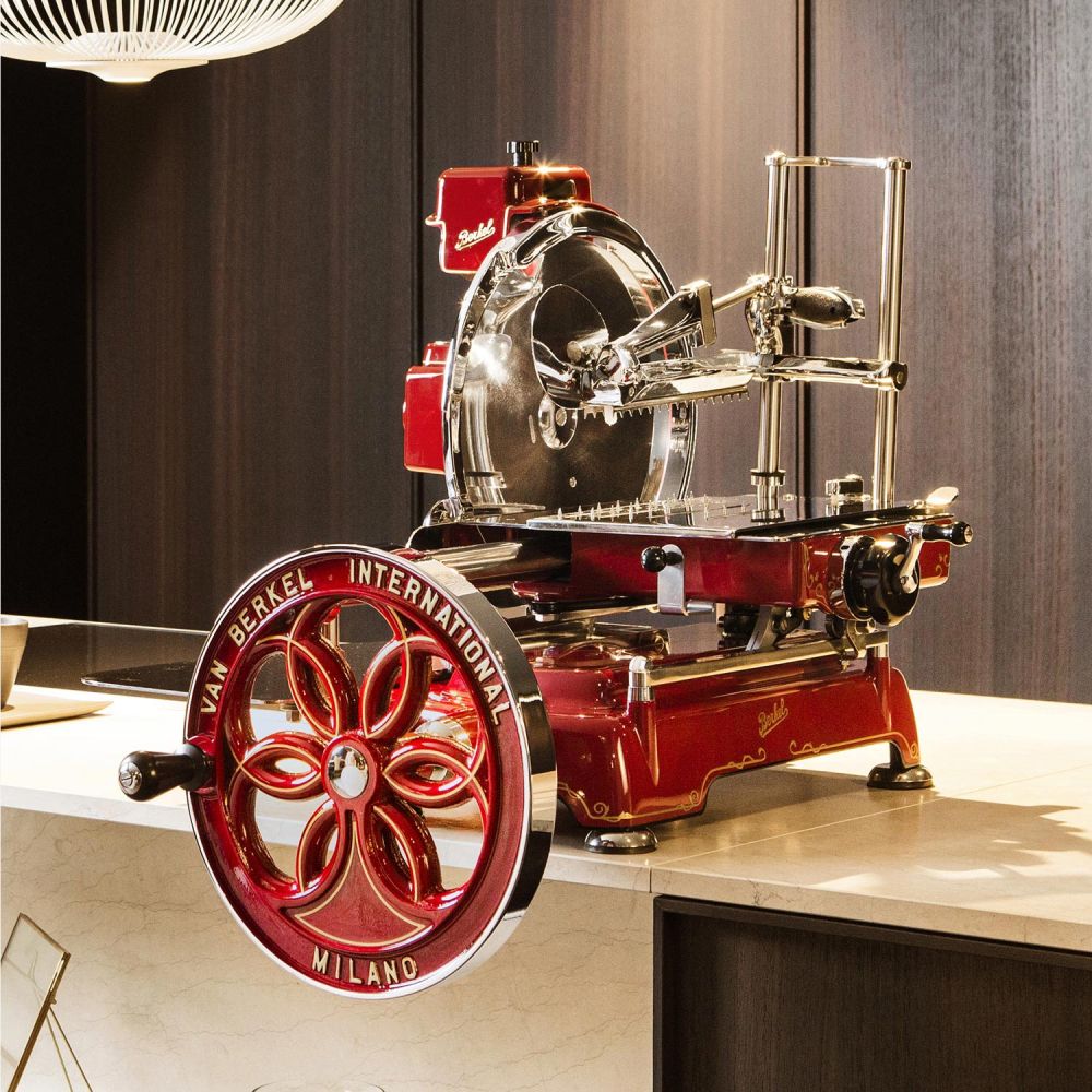 Eine Berkel Volano B114 Rot manuelle Aufschnittmaschine mit passgenauen Einlegebrett, die ihre aufwendige Metallverarbeitung und ein großes Rad mit der Aufschrift „Van Berkel International Milano“ zur Schau stellt, steht prominent auf einer Marmorarbeitsplatte in einer modernen Küche.
