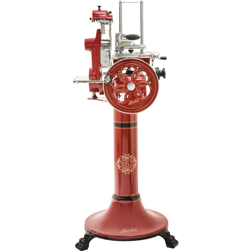 Ein rot-silberner Fleischschneider im Vintage-Stil, montiert auf einem passenden Sockel. Diese Berkel Volano B2 Rot Schwungradmaschine - Wunderschöne Aufschnittmaschine + Fassholzbrett Unikat verfügt über ein handgekurbeltes Rad mit aufwendigen Details und kunstvollen goldenen Akzenten. Der Sockel hat dekorative Elemente und steht auf schwarzen, verzierten Beinen.