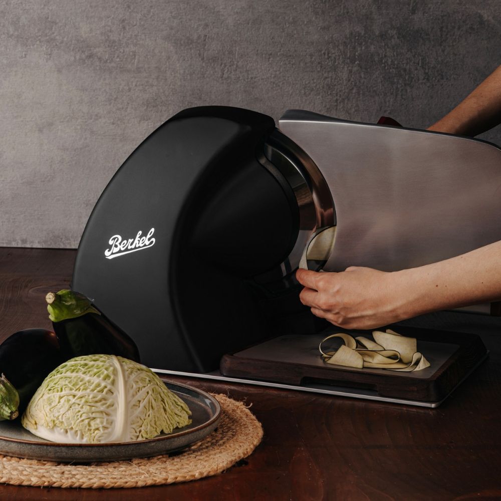 Eine Person schneidet Zucchini mit der Berkel Home Line 250 Plus schwarz matt Aufschnittmaschine mit handgefertigtem Schneidebrett & Wellenschliff-Bausatz für Brot auf einem Holztisch, neben Kohl und zwei Auberginen.