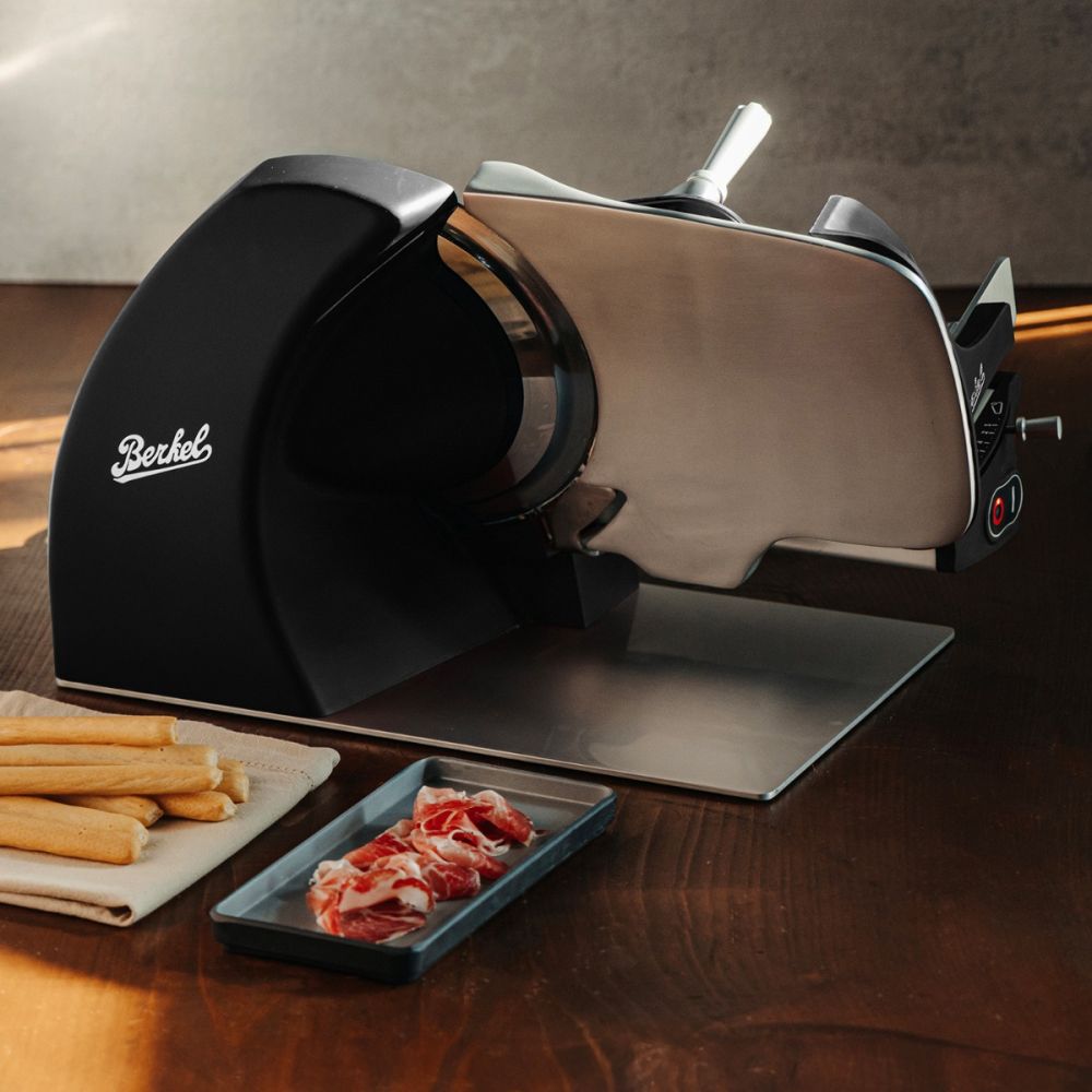 Die Berkel Home Line 250 Plus schwarz matt - Aufschnittmaschine neues Modell mit handgefertigtem Unikat-Schneidebrett & Wellenschliff-Bausatz für Brot steht auf einem Holztisch, umgeben von Brotscheiben und Brotsticks bei warmem Licht.