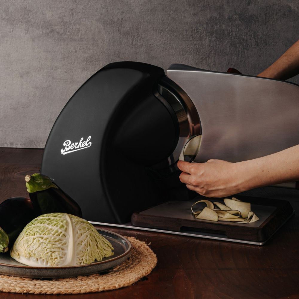 Eine Person schneidet mit der Berkel Home Line 200 Plus schwarz matt Aufschnittmaschine Gemüse auf einem handgefertigten Schneidebrett, während Kohl und Auberginen auf einer geflochtenen Matte daneben liegen.