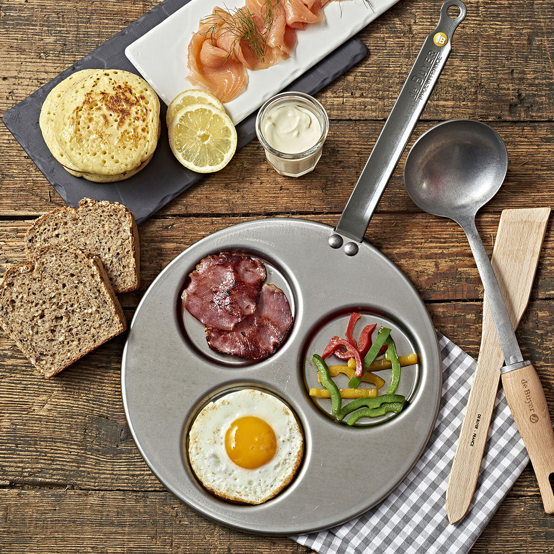 Eine dreiteilige De Buyer Blinis-Pfanne Mineral B enthält ein Spiegelei, knusprigen Speck und sautierte Paprika. Daneben liegen Roggenbrotscheiben, ein englischer Muffin, Räucherlachs, Zitrone und ein Glas Sauce auf einem rustikalen Holztisch.