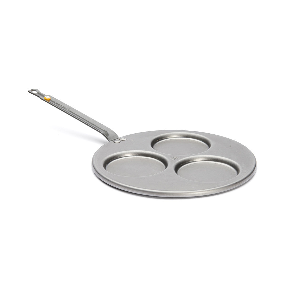 Die De Buyer Blinis-Pfanne Mineral B ist eine Metallpfanne mit drei kreisförmigen Fächern und einem langen Stiel. Sie eignet sich perfekt, um mehrere kleine Speisen wie Eier, Pfannkuchen oder Blinis auf einmal zuzubereiten.