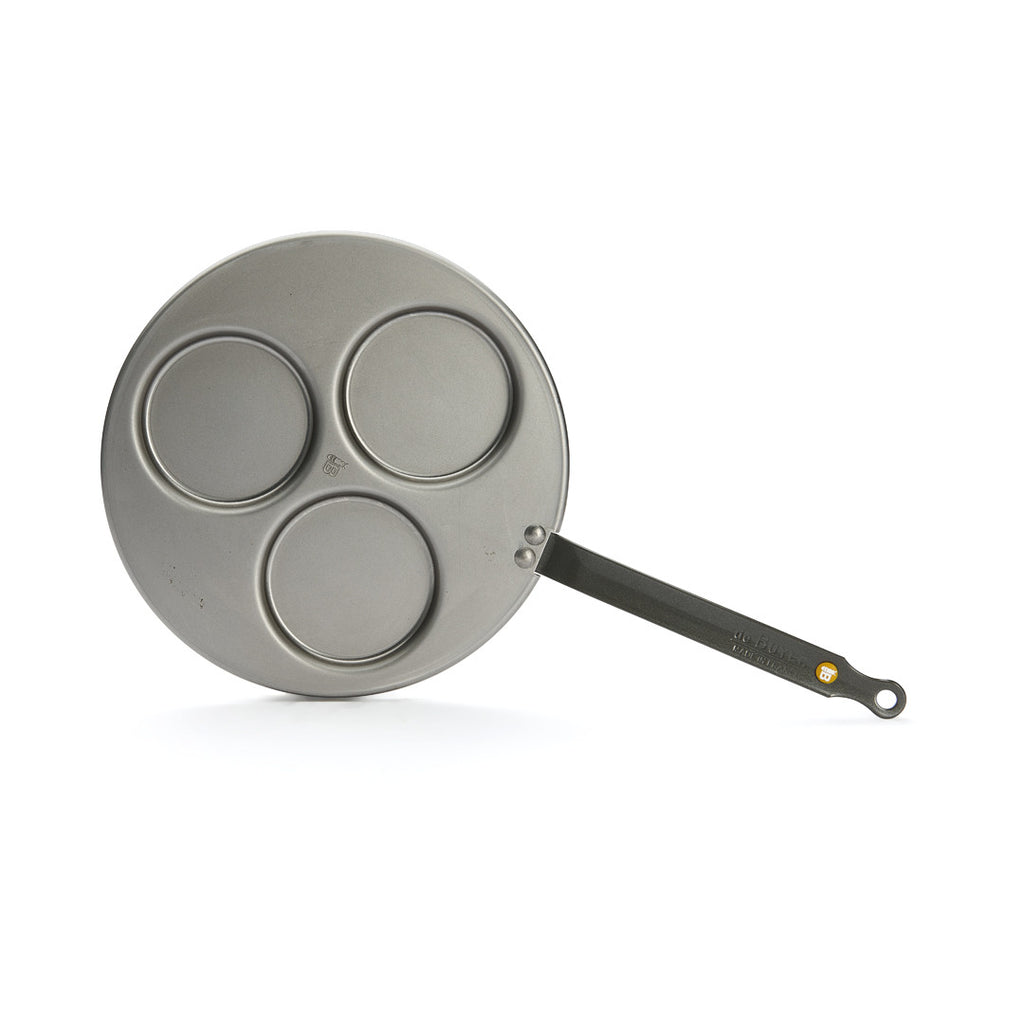 Die Blinis-Pfanne Mineral B von De Buyer ist eine Metallpfanne mit drei kreisförmigen Vertiefungen und einem langen schwarzen Griff. Sie eignet sich perfekt für die Zubereitung mehrerer Blinis auf einmal. Sie ist vor einem weißen Hintergrund fotografiert.