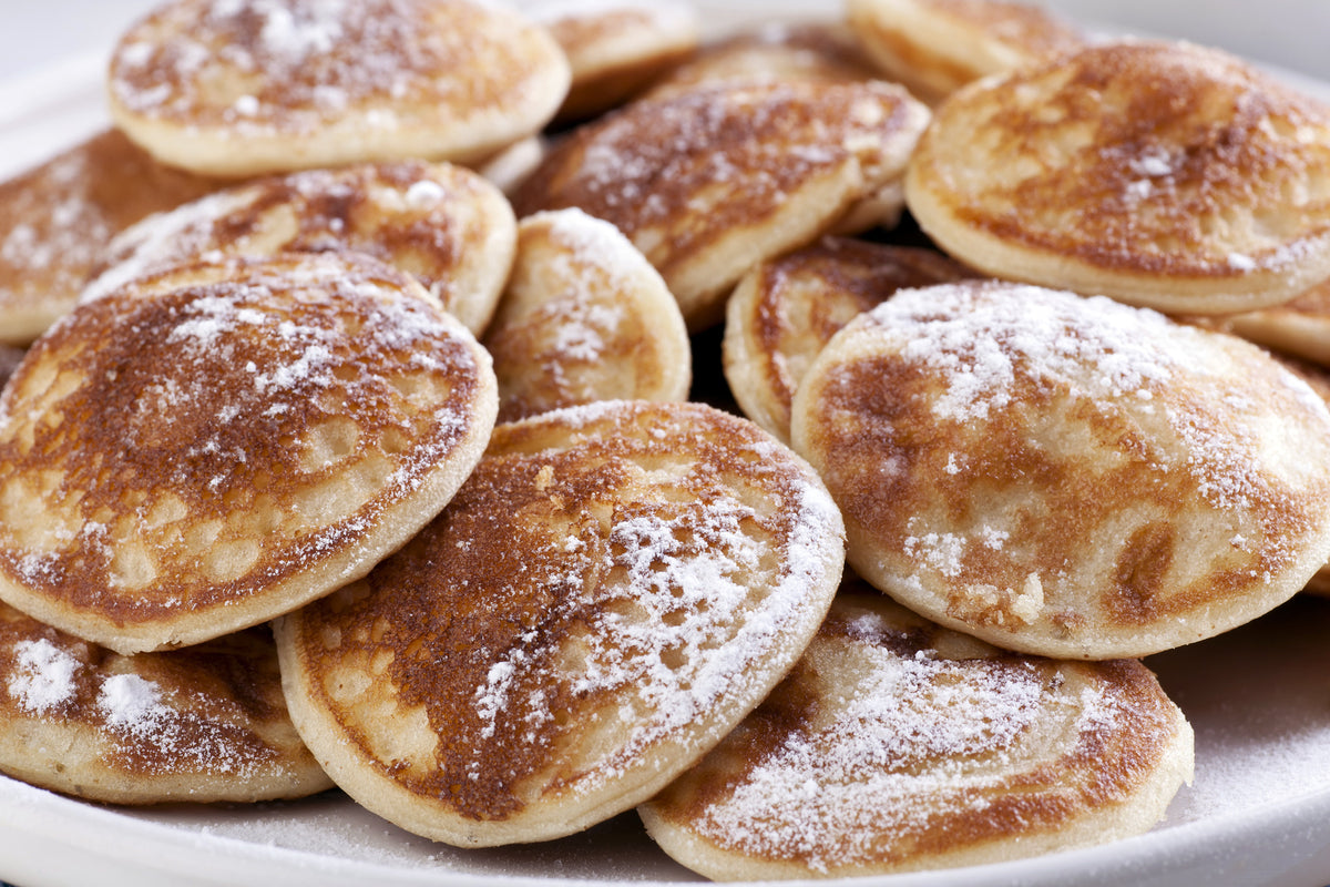 Nahaufnahme eines Tellers mit kleinen, goldbraunen, mit Puderzucker bestäubten Pfannkuchen, frisch zubereitet mit der De Buyer Blinis-Pfanne Mineral B.