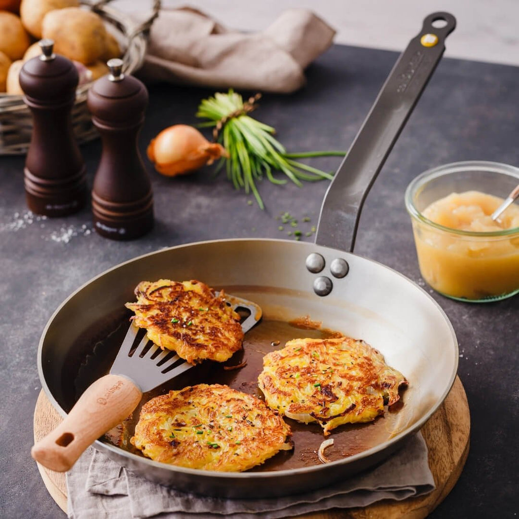 Auf der De Buyer Bratpfanne Mineral B liegen drei goldbraune Kartoffelpuffer, einer davon auf einem Spatel. Daneben stehen Apfelmus, Zwiebeln, Schnittlauch, ein Korb mit Kartoffeln und zwei hölzerne Pfeffermühlen auf einer dunklen Arbeitsplatte.
