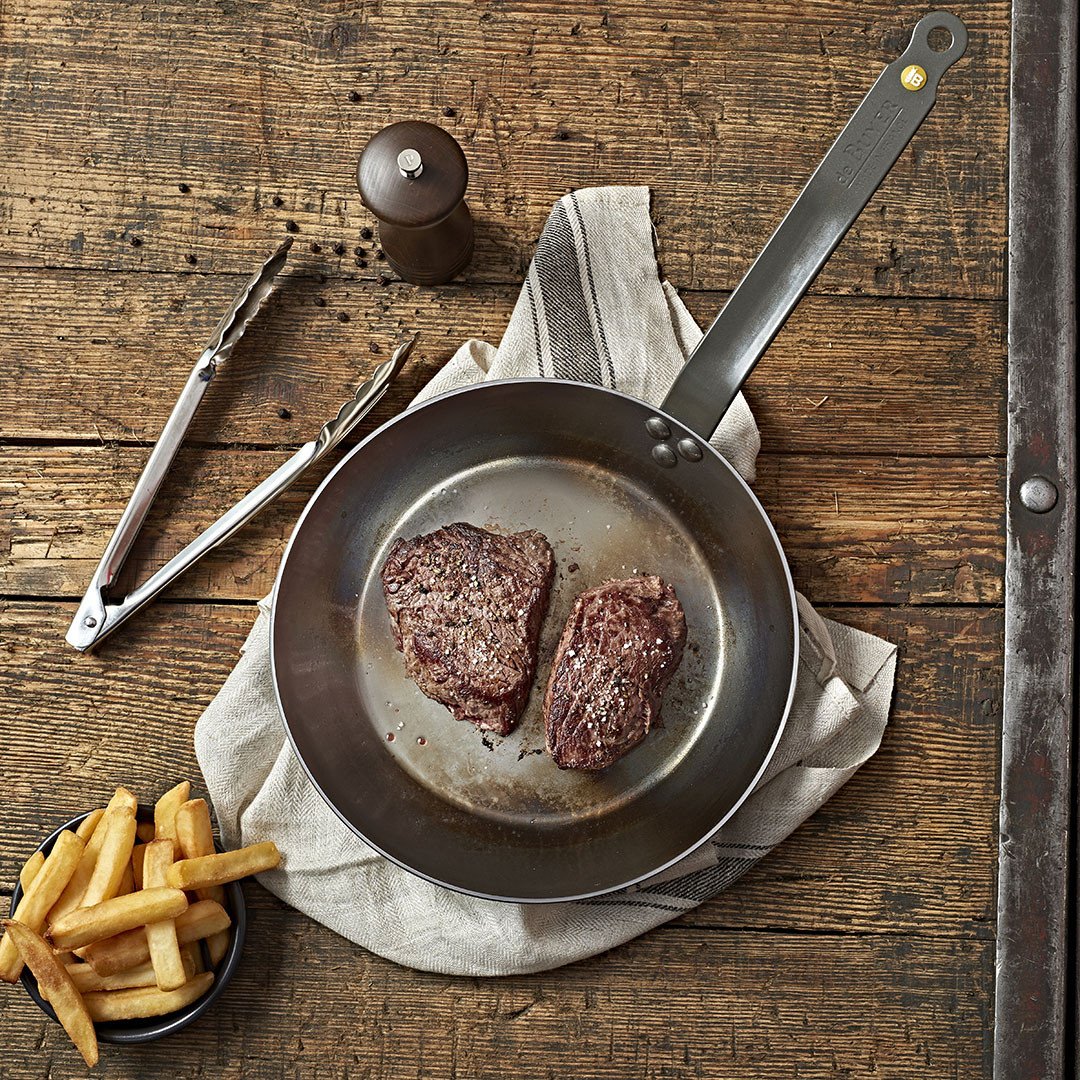 Die De Buyer Bratpfanne Mineral B mit zwei saftigen Steaks steht auf einem Tuch auf rustikalem Holztisch, daneben eine Schale Pommes, eine Grillzange und eine Pfeffermühle.