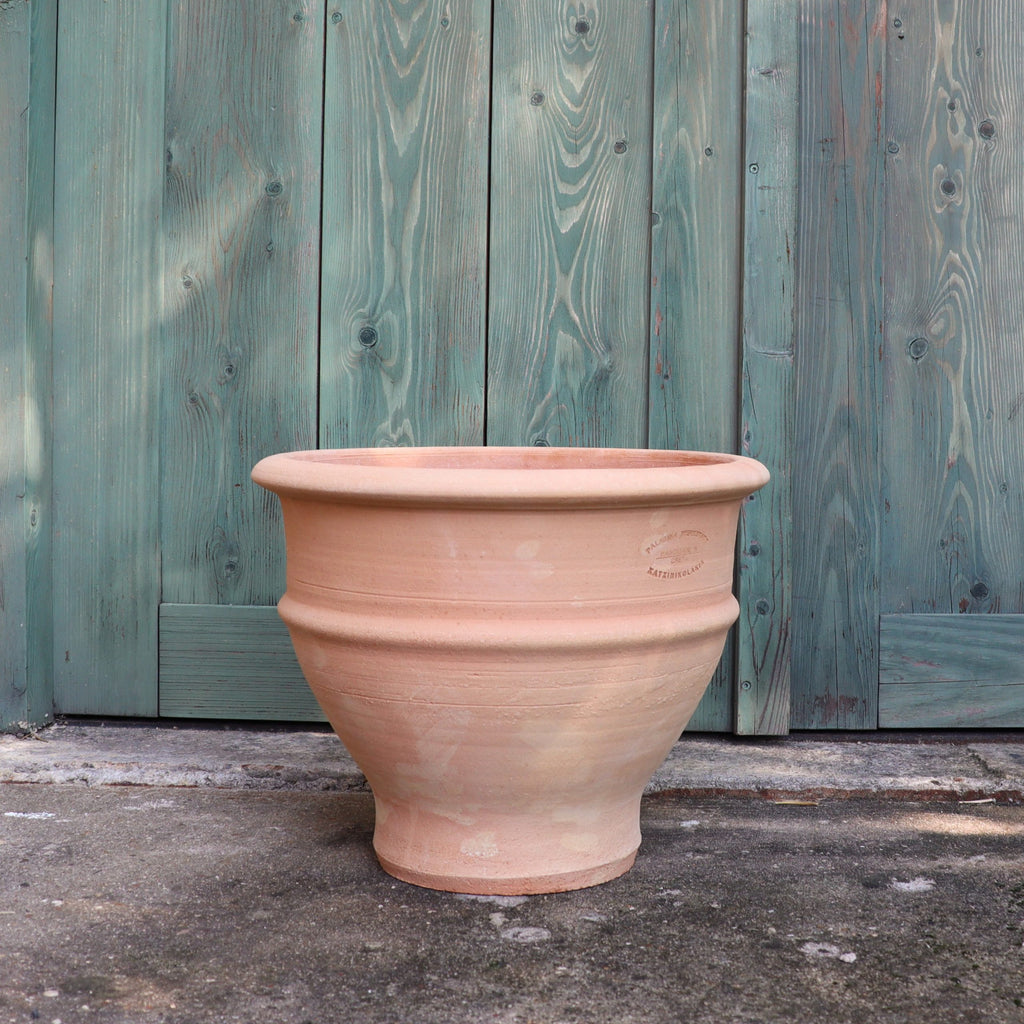 Das frostfeste Ton-Pflanzgefäß "Buxus", 40 cm, steht leer auf einer Betonfläche vor einer verwitterten tealfarbenen Holztür mit vertikalen Paneelen.