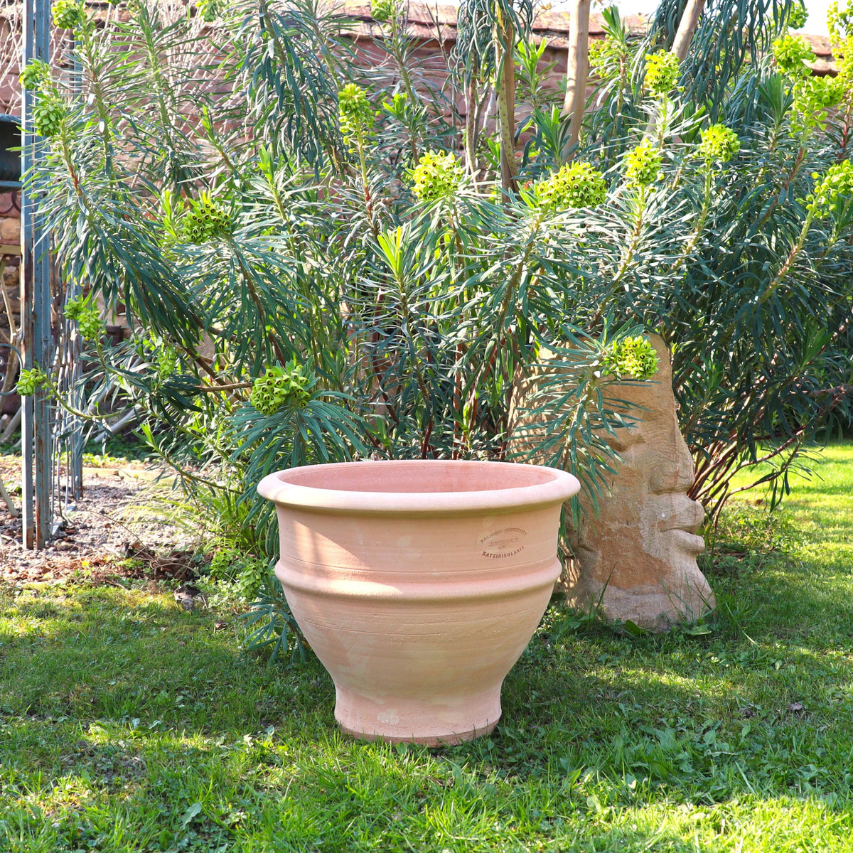 Das frostfeste Ton-Pflanzgefäß "Buxus", 40 cm, steht auf grünem Gras zwischen üppigen hohen Pflanzen und hellem Laub in einem sonnenbeschienenen Garten, mit einer dekorativen Steinskulptur, die teilweise hinter dem Topf sichtbar ist.