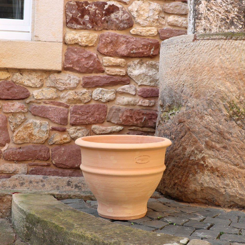 Das frostfeste Ton-Pflanzgefäß "Buxus", 40 cm, steht leer auf Kopfsteinpflaster neben einer Mauer aus unregelmäßigen roten und beigen Steinen. Rechts ist ein Teil einer alten Steinstruktur zu sehen.