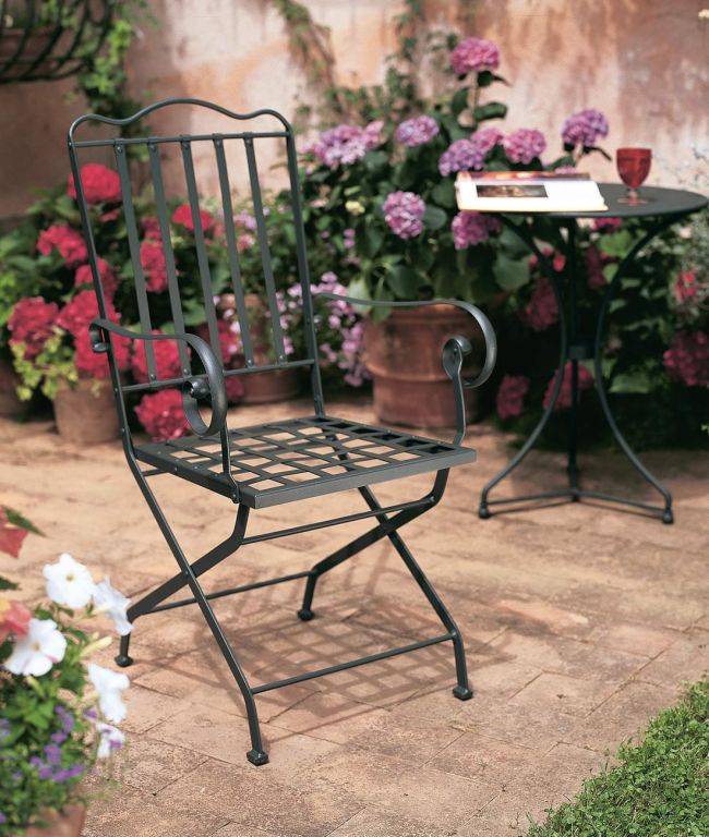 Ein klappbarer Stuhl Toscana mit Gittersitz steht auf einer gemauerten Terrasse. Daneben steht ein kleiner runder Tisch mit einem Buch und einem Glas. Hinter dem Stuhl stehen Topfpflanzen und blühende Blumen in verschiedenen Farben, die der ruhigen Gartenumgebung einen Farbtupfer verleihen.