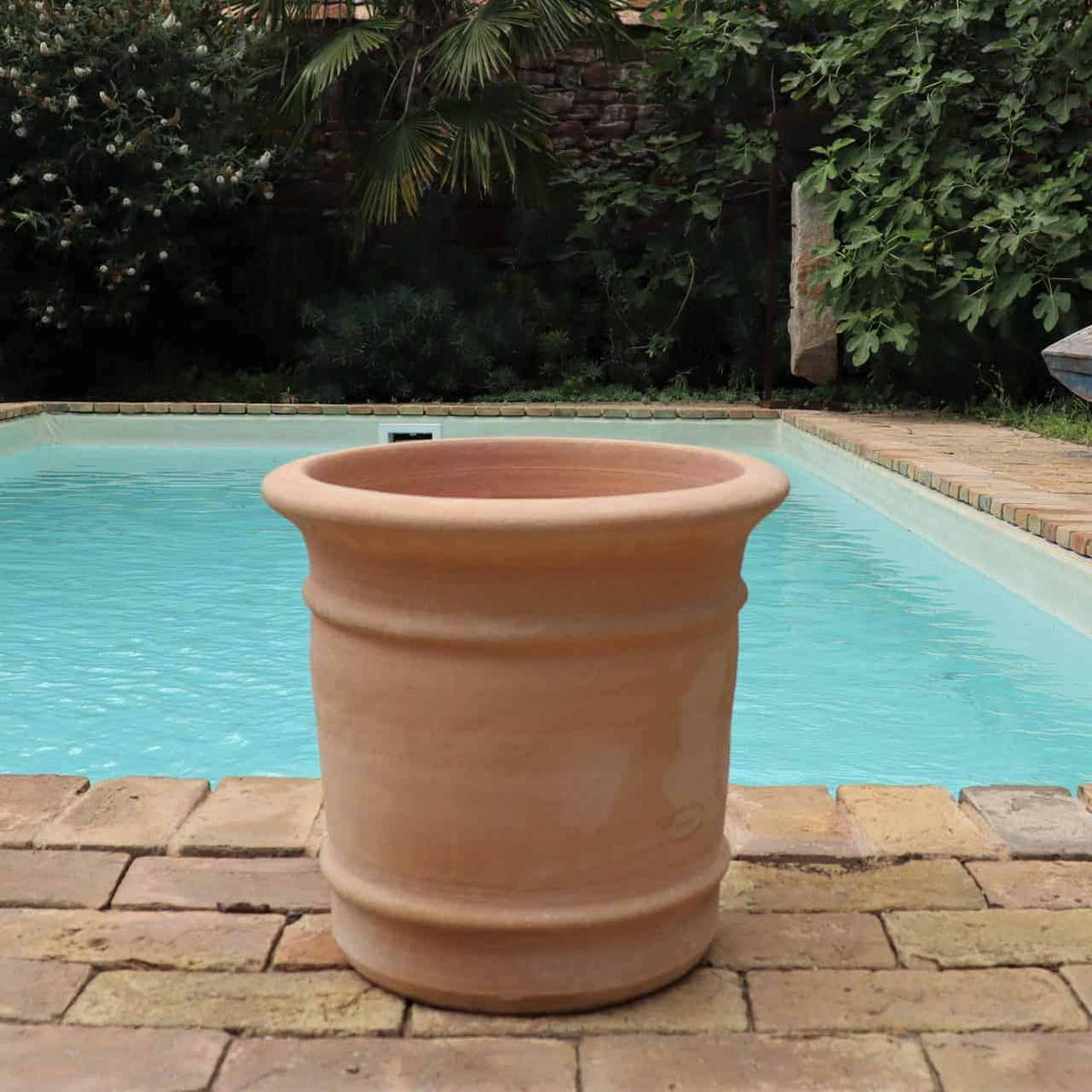 Ein großer, leerer Terrakotta-Pflanzkübel „Canna“ H=33 cm steht auf einer gemauerten Terrasse vor einem klaren, blauen Swimmingpool. Üppiges Grün, darunter Bäume und Büsche, bilden eine ruhige Kulisse für die Szene. Die Atmosphäre ist ruhig und einladend, perfekt zum Pflanzen leuchtender Canna-Blüten.