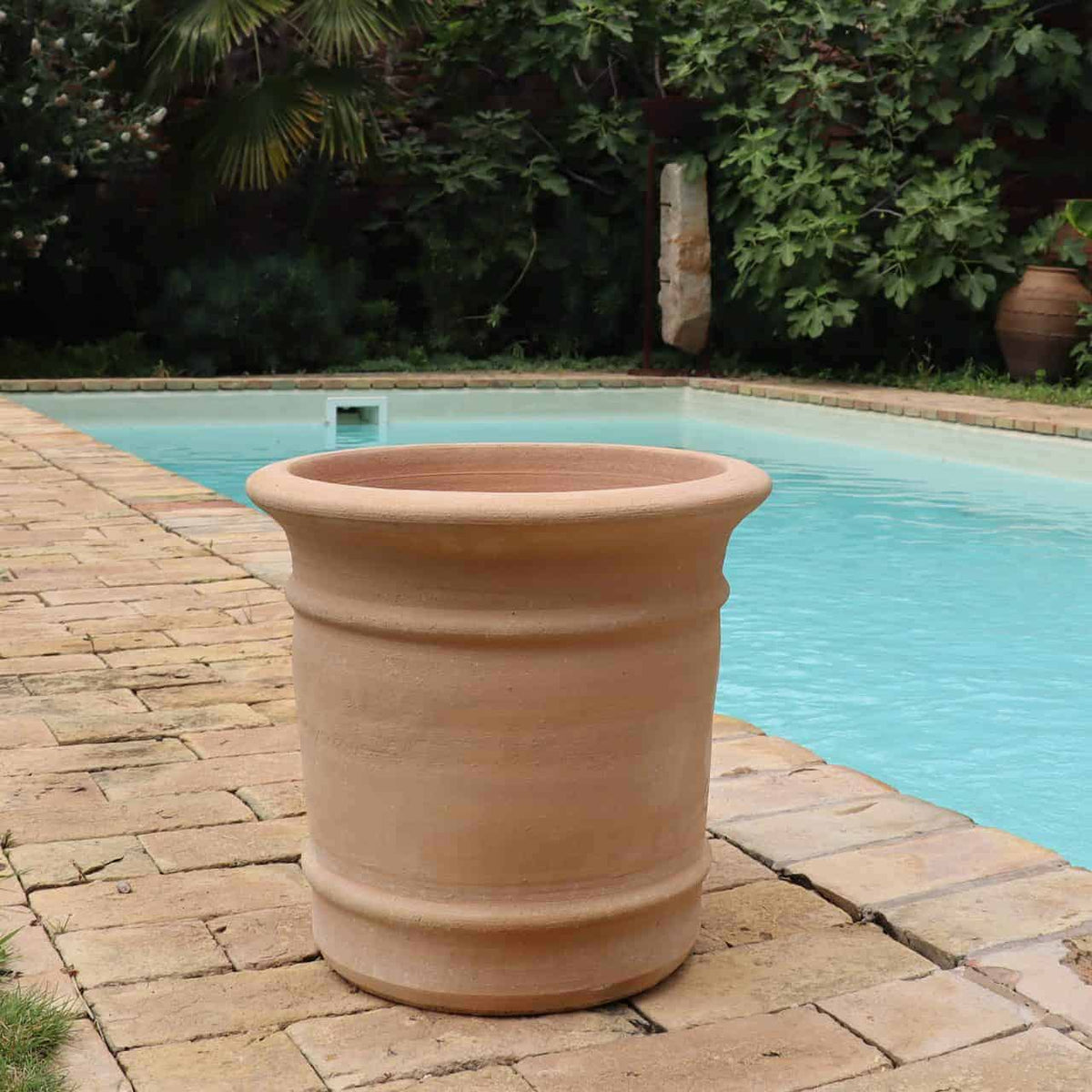 Ein großer, leerer Terrakotta-Pflanzkübel „Canna“ H=33 cm steht auf einem mit Steinen gepflasterten Pooldeck. Der Pool im Hintergrund ist mit klarem, blauem Wasser gefüllt und üppiges grünes Laub umgibt den Bereich.