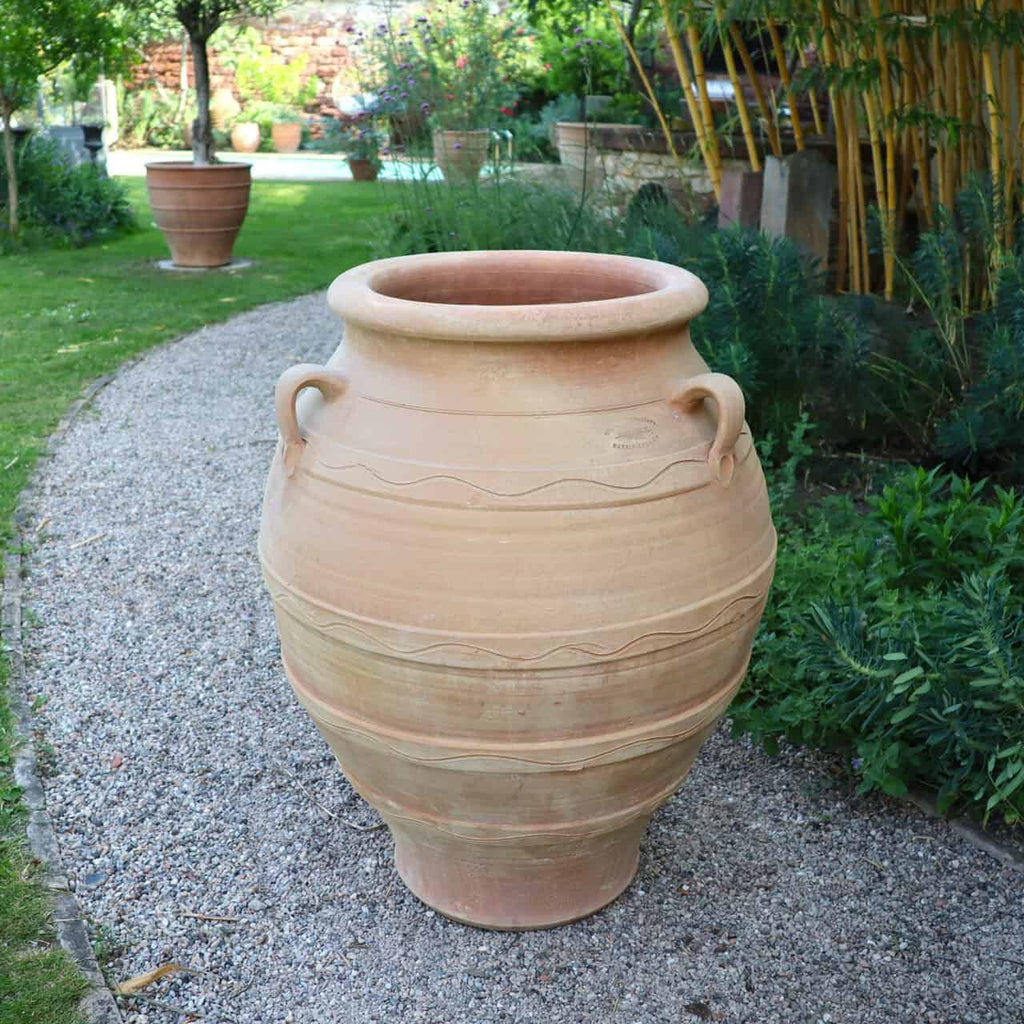 Die winterfeste Dekorationsamphore "Cassia", H=70 cm, mit Henkeln steht auf einem Kiesweg im Garten, umgeben von grünen Pflanzen und Bambus; im Hintergrund sind weitere Töpfe und üppiges Grün zu sehen.
