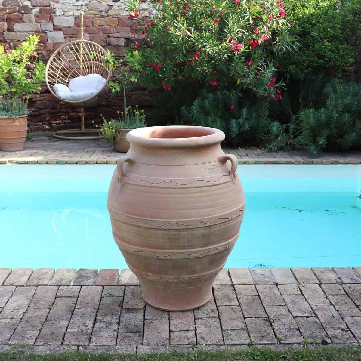 Die winterfeste Dekorationsamphore "Cassia", H=70 cm, steht auf einer Ziegelterrasse vor einem türkisfarbenen Pool, daneben ein Korbstuhl und üppige grüne Pflanzen.