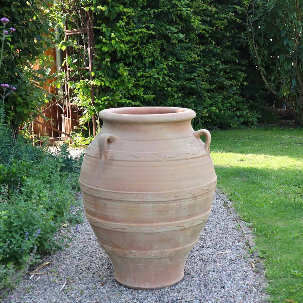 Eine winterfeste Dekorationsamphore "Cassia", H=70 cm, mit Henkeln steht auf einem Kiesweg im üppigen Garten und ist von viel Grün umgeben.