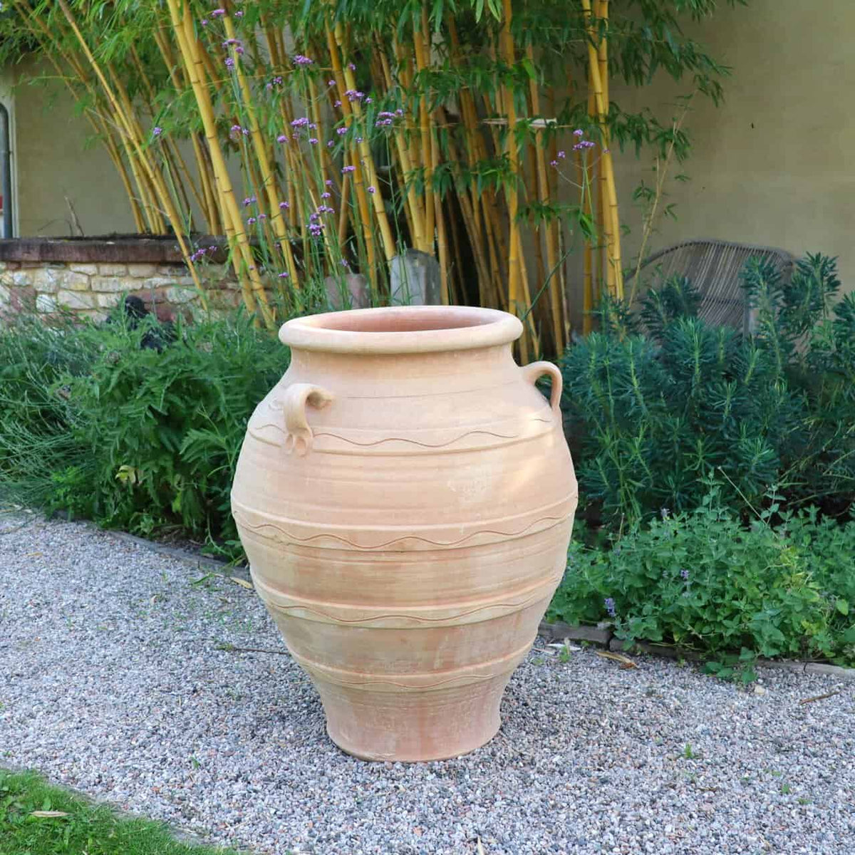 Der winterfeste Dekorationsamphore "Cassia", H=70 cm, mit Zierrippen und Griffen, steht auf einem Kiesweg in einem üppigen Garten mit Grünpflanzen, Bambus und einer gelben Wand im Hintergrund.