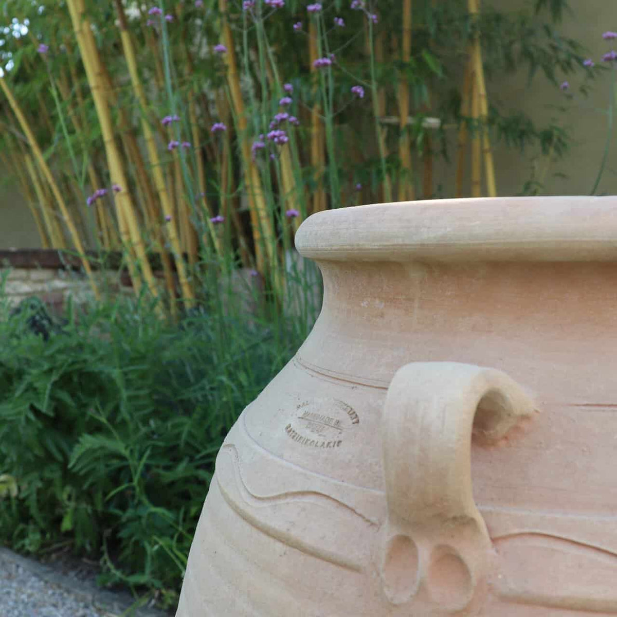 Die winterfeste Dekorationsamphore "Cassia", H=70 cm, mit geschwungenem Henkel steht im Garten, umgeben von Bambus, Kassia, violetten Blüten und einem Kiesweg am Sockel.
