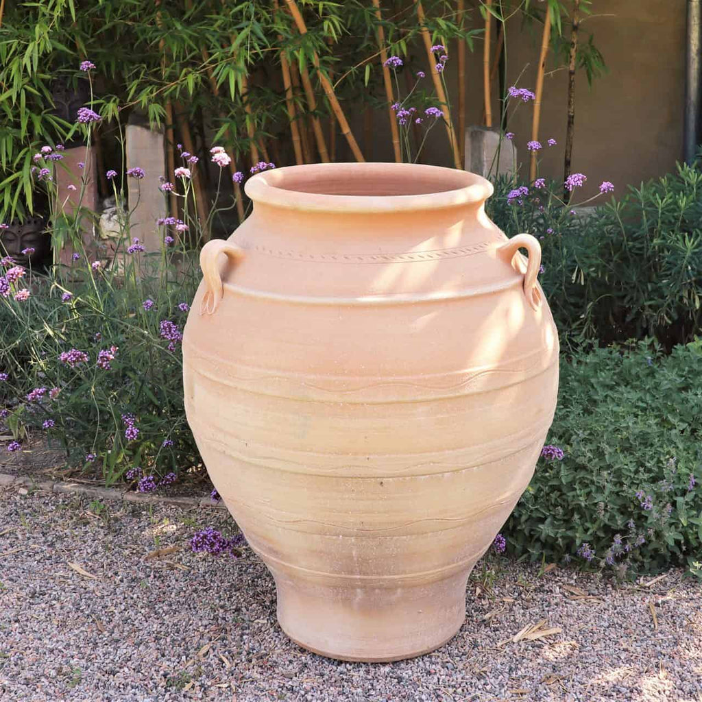 Ein großer, beiger Keramiktopf mit zwei Griffen steht auf einer Kiesfläche in einem Garten. Es handelt sich um eine frostfeste Amphore „Cassia“, H=100 cm, umgeben von grünen Sträuchern und zarten lila Blüten, im Hintergrund stehen hohe Bambuspflanzen. Das Sonnenlicht wirft weiche Schatten und hebt die glatten, abgerundeten Konturen des Topfes hervor.