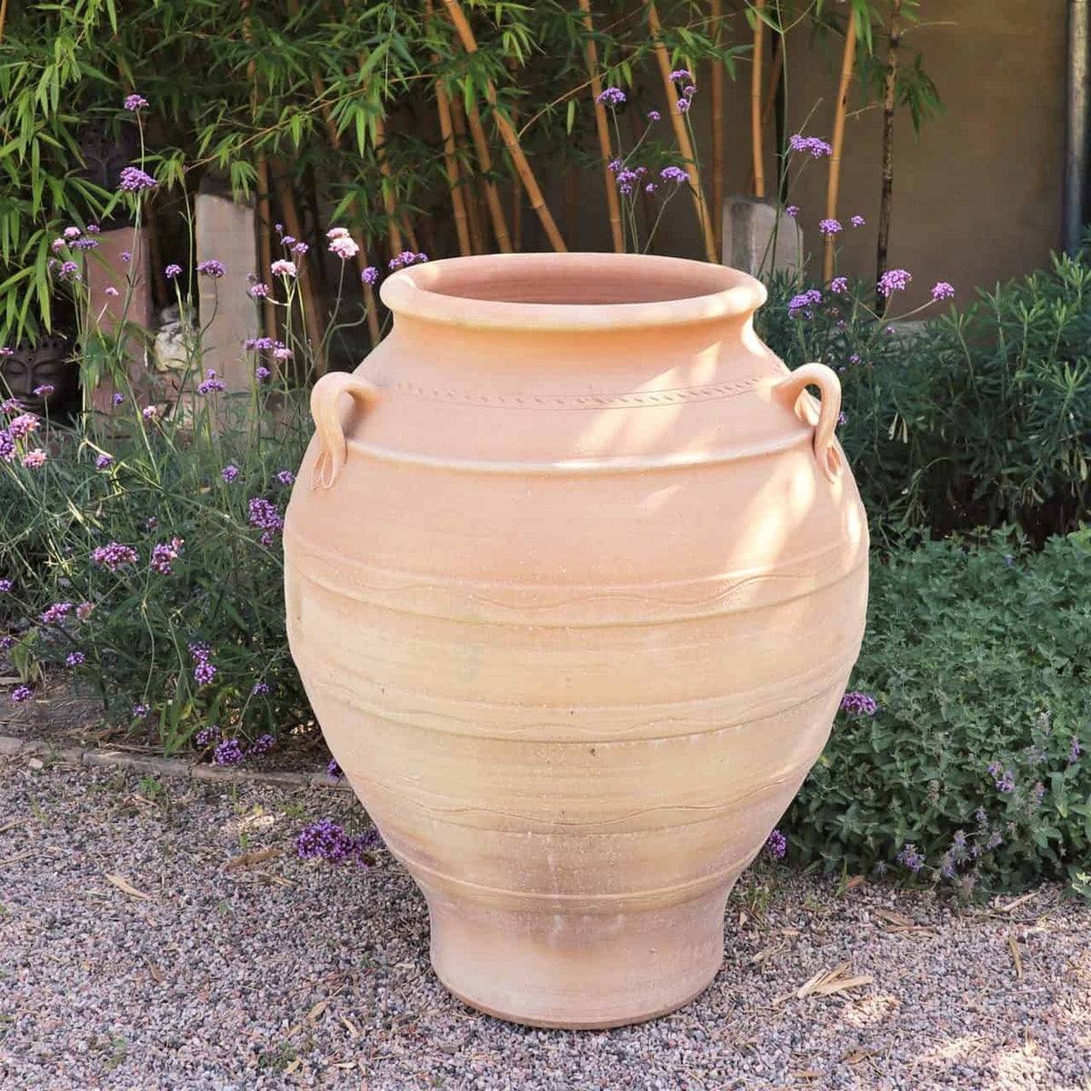 Ein großer, beiger Keramiktopf mit zwei Griffen steht auf einer Kiesfläche in einem Garten. Es handelt sich um eine frostfeste Amphore „Cassia“, H=100 cm, umgeben von grünen Sträuchern und zarten lila Blüten, im Hintergrund stehen hohe Bambuspflanzen. Das Sonnenlicht wirft weiche Schatten und hebt die glatten, abgerundeten Konturen des Topfes hervor.