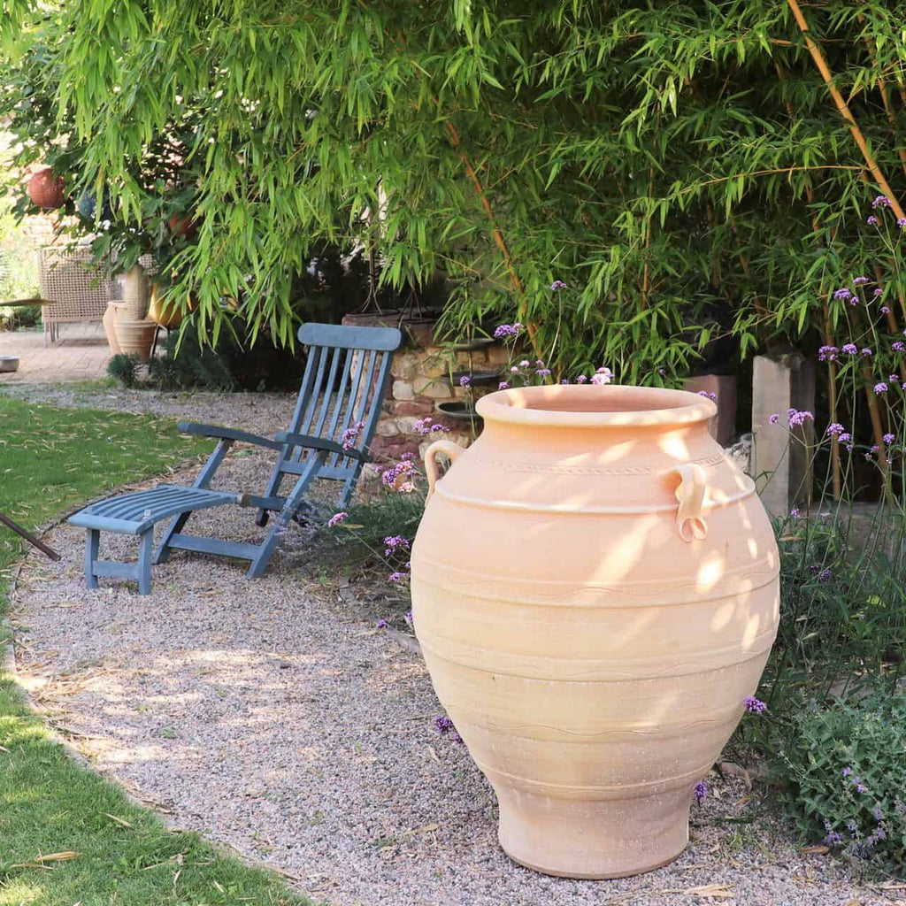 Eine ruhige Gartenszene mit der großen, frostfesten Amphore „Cassia“ (H=100 cm) mit Henkeln auf einem Kiesweg. Daneben stehen zwei blaue Holzliegestühle unter einer üppigen Bambuspflanze, umgeben von violetten Blüten und grünem Gras. Der Hintergrund enthält weitere Pflanzen und Gartendeko.