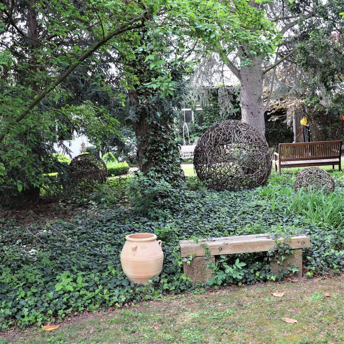 Eine ruhige Gartenszene zeigt eine Holzbank, kugelförmige Drahtskulpturen, üppiges Grün und einen mit Efeu bewachsenen Baum. Im Vordergrund sind die griechische Amphore „Cassia“ (H=50 cm) und eine Steinbank von Bodendeckerpflanzen umgeben.