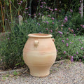 Eine große griechische Amphore aus beigefarbenem Ton „Cassia“ (H=50 cm), geschmückt mit zwei kleinen Henkeln, steht auf einem Kiesweg, umgeben von üppigen grünen Blättern und großen violetten Blüten vor einem Hintergrund aus verschiedenen Gartenpflanzen und hoch aufragenden Zweigen.