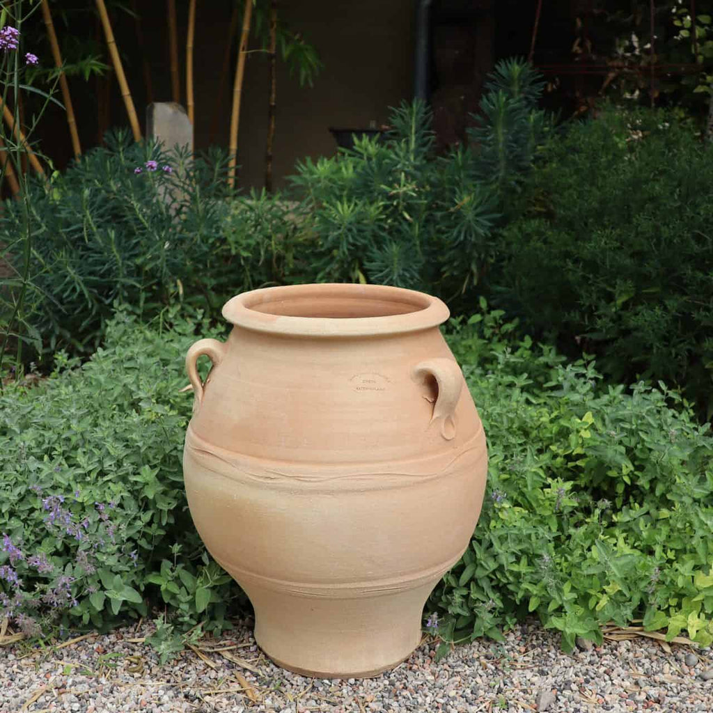 Die griechische Amphore „Cassia“, ein großer beigefarbener Tontopf mit zwei runden Griffen, steht auf einem Kiesweg inmitten üppiger Vegetation. Ihr schlichtes, rustikales Design steht im Mittelpunkt des Bildes.