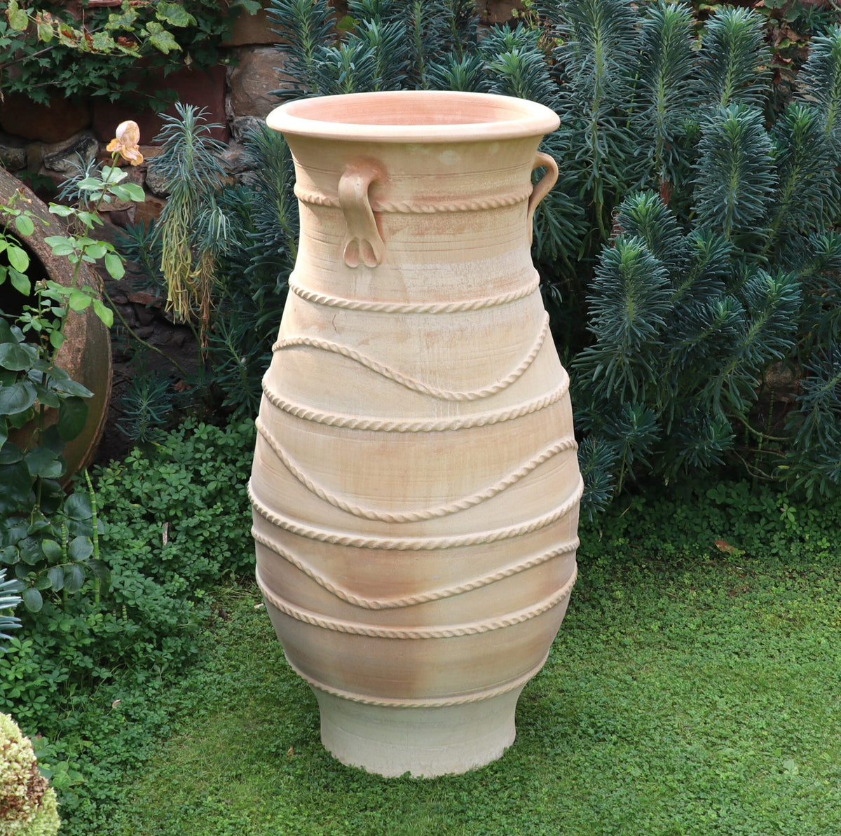 Die frostbeständige griechische Amphorenvase „Cistus“ (H=1,30m), eine große beige Keramikvase mit dekorativen horizontalen Rillen und zwei kleinen Henkeln oben, steht auf einer Rasenfläche vor einem Hintergrund aus üppigen grünen Pflanzen und Cistusblättern. Die Vase weist ein strukturiertes Wellenmuster auf, das ihren Körper umgibt.