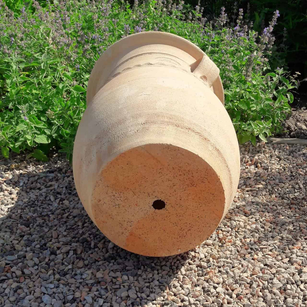 Die griechische Amphorenvase "Cistus", H=40cm, liegt auf der Seite auf Kies, mit grünen Blattpflanzen und lila Blüten im Hintergrund. Der Sockel ist mit einem zentralen Abflussloch versehen.