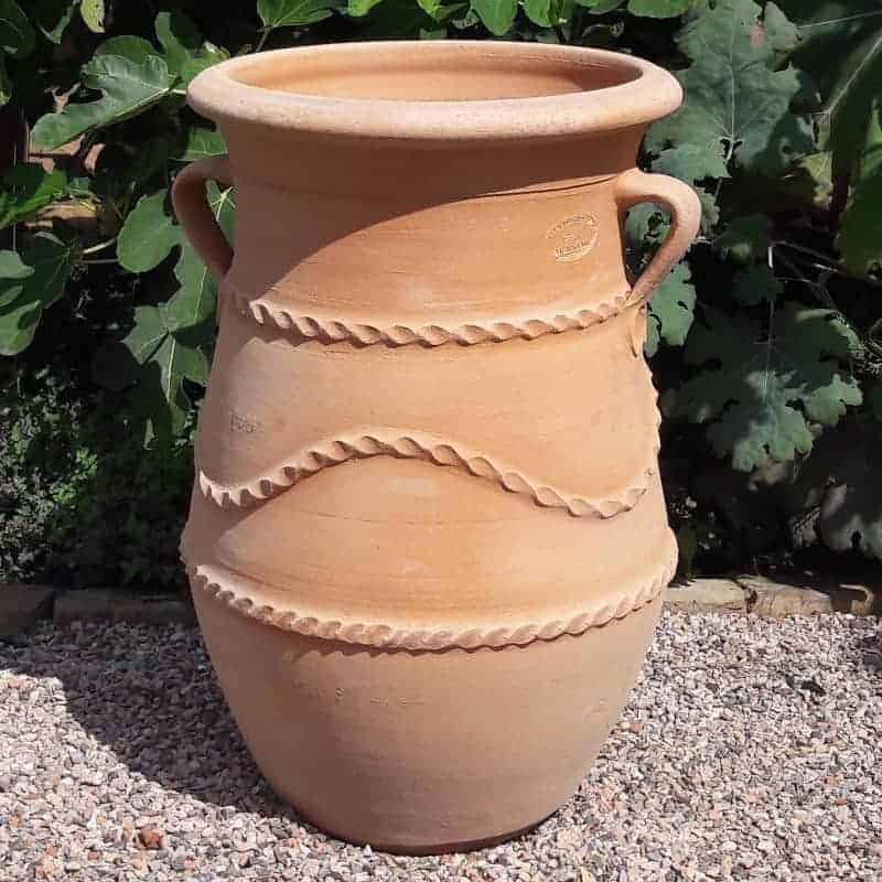 Eine große griechische schmale Terrakotta-Amphore „Cistus“ H=60 cm mit zwei Henkeln und dekorativen horizontalen Rillen steht auf einer Kiesfläche und erinnert an ein antikes Artefakt. Im Hintergrund ist üppiges grünes Laub zu sehen.