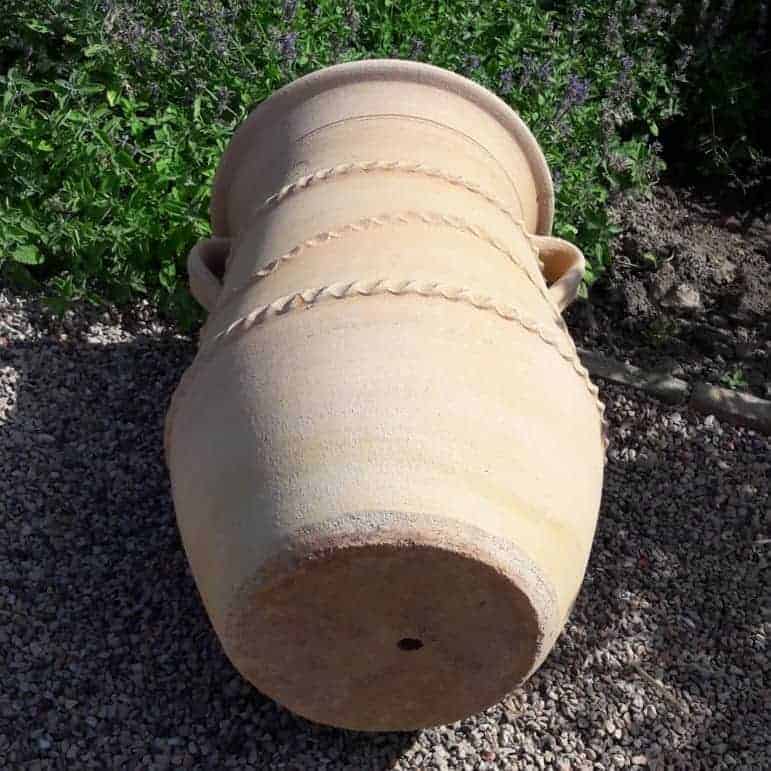 Die griechische schmale Terrakotta-Amphore „Cistus“ H=60 cm, eine große, hellbeige Keramikurne, verziert mit dekorativen Bändern und zwei Henkeln, liegt auf der Seite auf einer Kiesfläche, vor einer Gartenkulisse mit üppigen grünen Blattpflanzen.