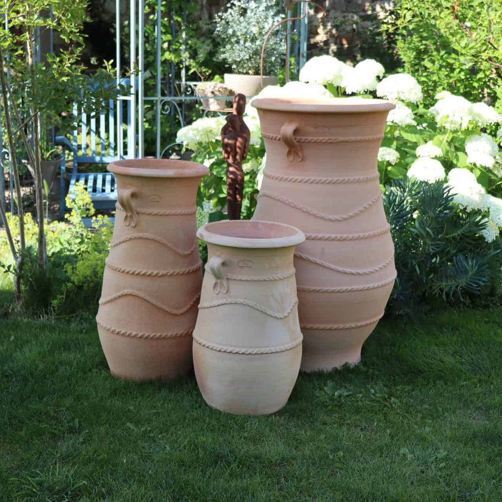 Drei griechische Amphorenvasen „Cistus“, jeweils 1 Meter hoch, sind in einem üppigen Garten angeordnet. Die Terrakotta-Pflanzgefäße sind mit dekorativen Seilen geschmückt und von Grünpflanzen umgeben, darunter weiße Hortensien und Cistus-Pflanzen. Im Hintergrund ist eine Skulptur einer menschlichen Figur zu sehen.