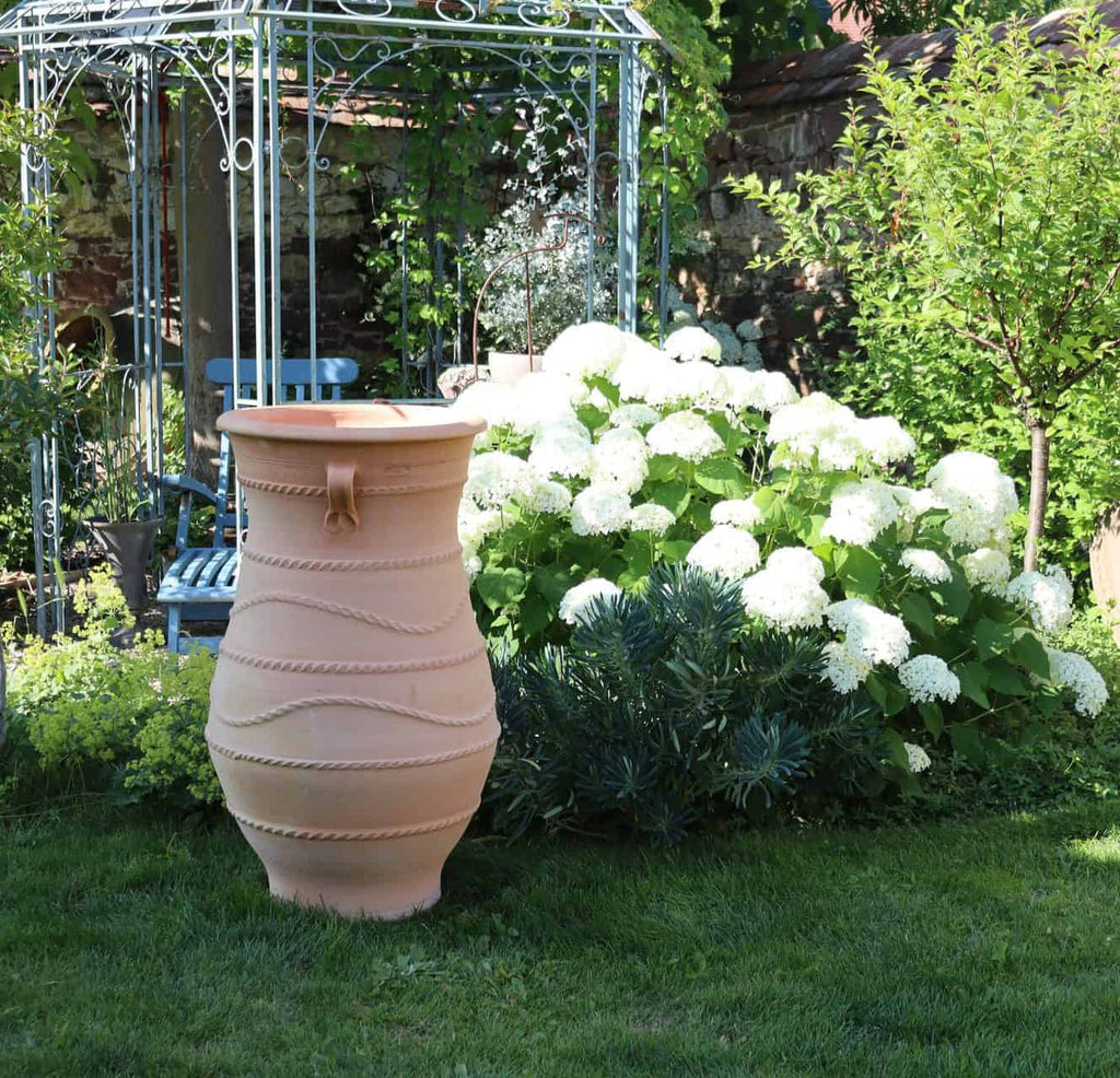 Eine terrakottafarbene Regentonne mit einem Hahn am Boden steht auf einer grünen Wiese neben einem blühenden Hortensienbusch. Daneben thront elegant eine griechische Amphorenvase „Cistus“ H=1m. Im Hintergrund sind ein metallischer Gartenpavillon und einige blaue Gartenmöbel inmitten üppigen Grüns zu sehen.