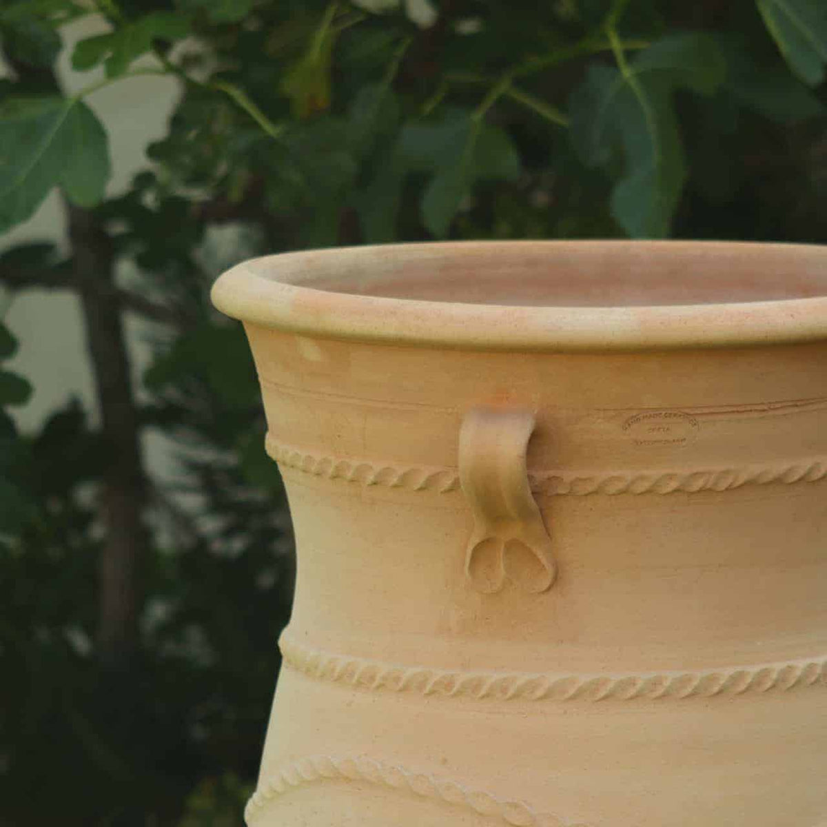 Eine Nahaufnahme einer griechischen Amphorenvase „Cistus“ H=1m mit einem strukturierten Design, das horizontale Reihen gedrehter Muster aufweist. Der 1 Meter hohe Topf steht vor einem Hintergrund aus üppigem grünem Laub.