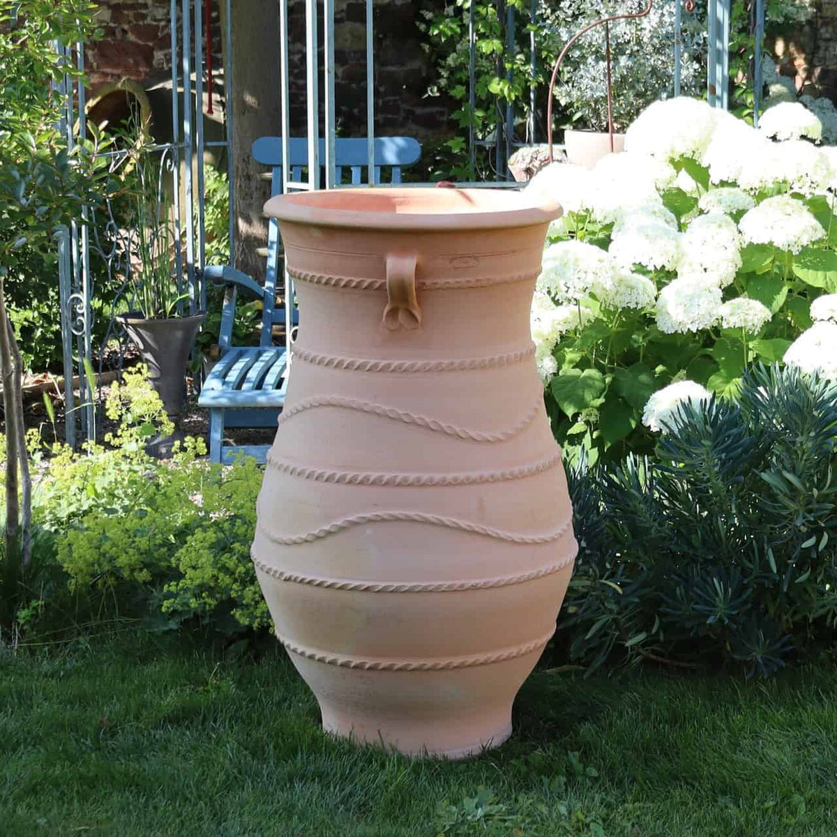 Eine griechische Amphorenvase „Cistus“ H=1m mit strukturiertem Seildesign steht auf einer Rasenfläche in einem Garten, umgeben von grünen Sträuchern, weißen Hortensien und verschiedenen Pflanzen. Im Hintergrund sind metallene Gartenstrukturen und ein blauer Stuhl zu sehen. Diese elegante Vase verleiht der Szene einen zusätzlichen Hauch von Charme.