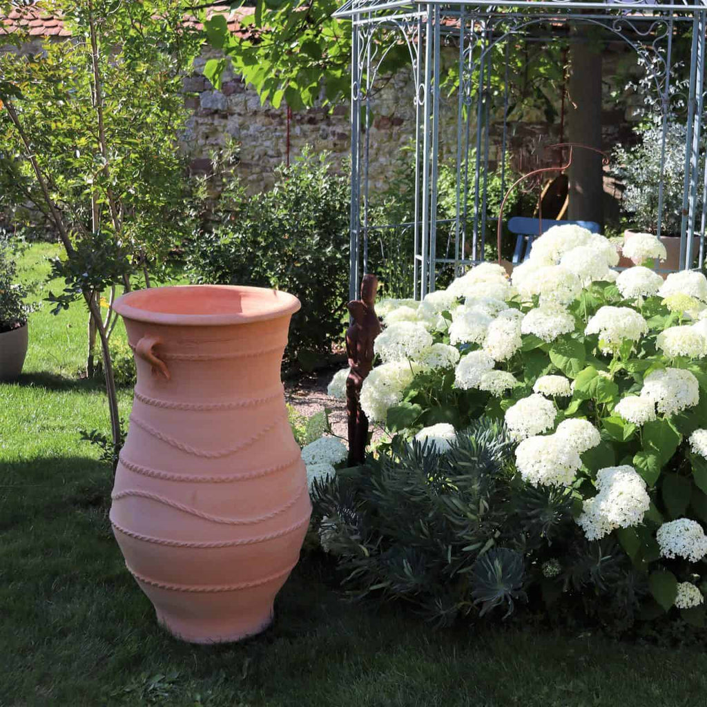 Eine große griechische Amphorenvase „Cistus“ H=1m steht auf einem üppigen grünen Rasen neben einem Gartenbeet mit blühenden weißen Cistus und verschiedenen Pflanzen. Im Hintergrund steht ein metallener Gartenpavillon, teilweise im Schatten von Bäumen.