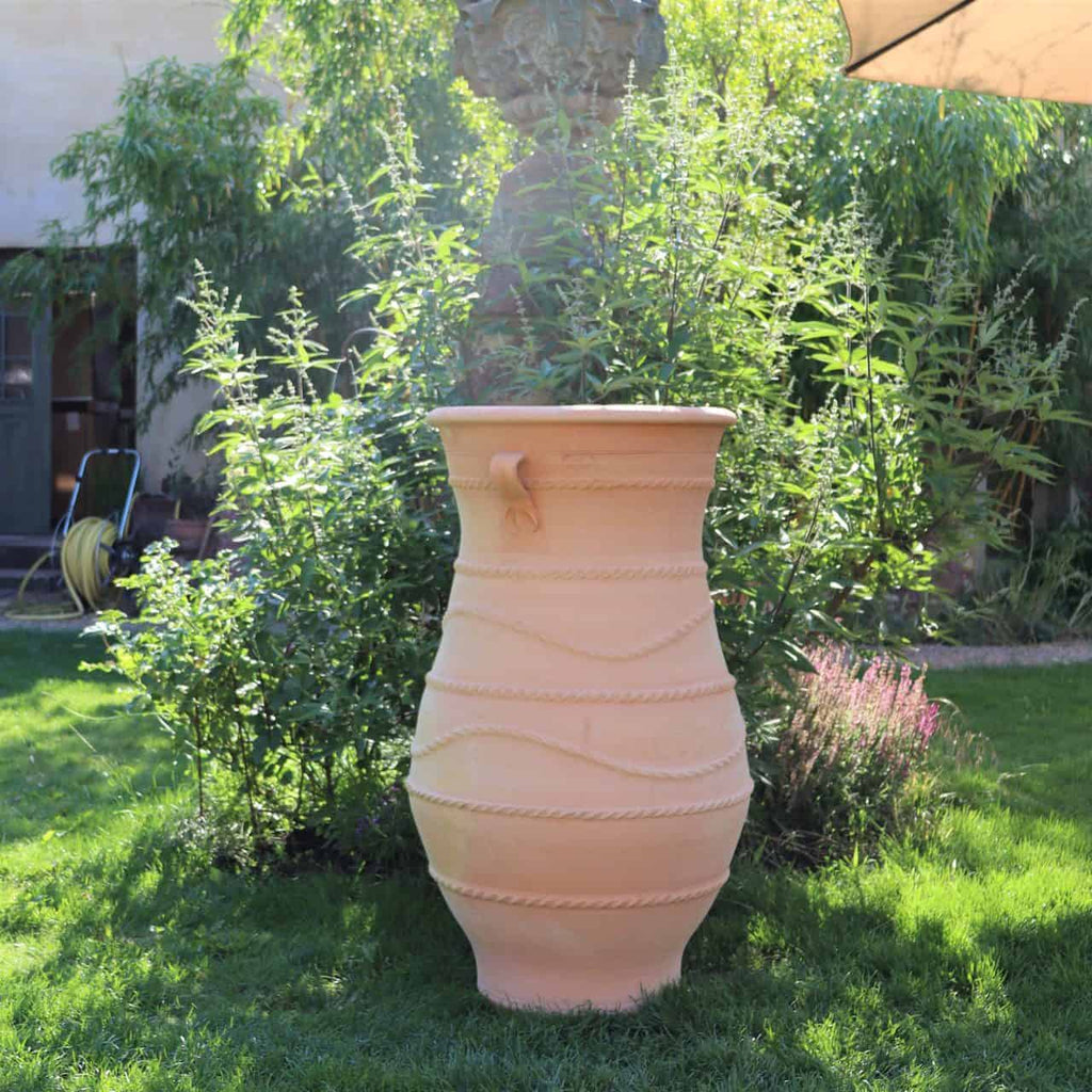 Eine große griechische Amphorenvase „Cistus“ H=1m mit aufwendigen Mustern steht auf einem üppigen grünen Rasen. Um den Rasen herum stehen dichte, belaubte Büsche und Bäume. Das Sonnenlicht erhellt die Szene und wirft weiche Schatten, und in der oberen rechten Ecke erstreckt sich ein teilweise sichtbarer Schirm.