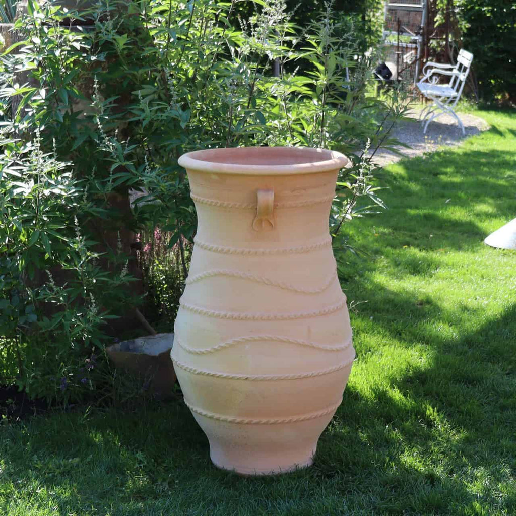 Eine große, beige griechische Amphorenvase „Cistus“ H=1m, geschmückt mit dekorativen horizontalen Bändern und einem einzelnen Griff, steht auf einer Rasenfläche. Sie ist von hohen, blattreichen Pflanzen umgeben, die an Cistus erinnern. Im Hintergrund ist ein weißer Metallstuhl zu sehen, was darauf hindeutet, dass es sich um einen Garten oder Hinterhof handelt.