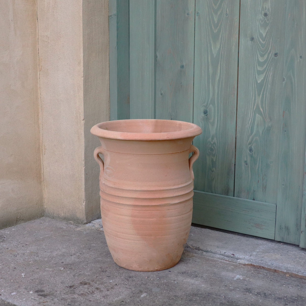Die hohe, leere Terrakotta-Vase "Cistus 2", H=40cm, mit zwei kleinen Henkeln steht auf einer Betonfläche vor einer grünen Holztür und beiger Wand.