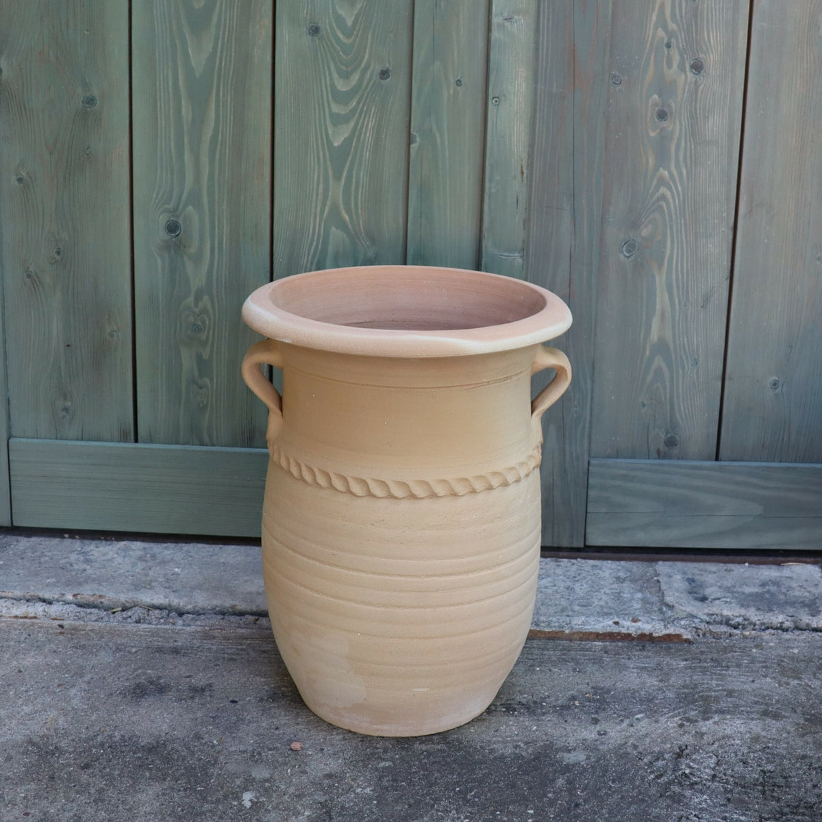 Eine große, beige griechische Amphorenvase „Cistus“ aus Keramik mit zwei Griffen und dekorativen Rillen in der Mitte steht auf einem Betonboden vor einem rustikalen grünen Holzzaun. Die 40 cm hohe Vase hat eine offene Oberseite und eine strukturierte, unglasierte Oberfläche.