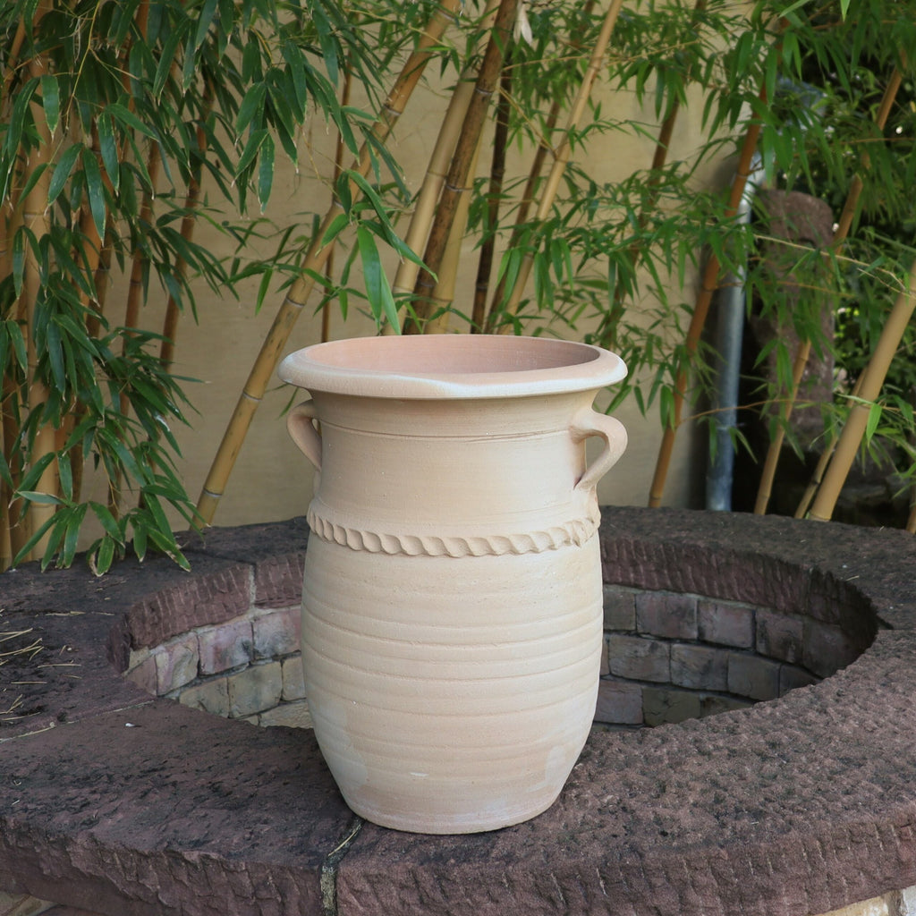 Eine große, beige griechische Amphorenvase „Cistus“ mit zwei Henkeln steht auf einer runden Steinstruktur. Grüne Bambuspflanzen bilden einen natürlichen Hintergrund. Die 40 cm große Vase hat ein schlichtes, rustikales Design mit einem seilartigen Detail um ihren Mittelteil.