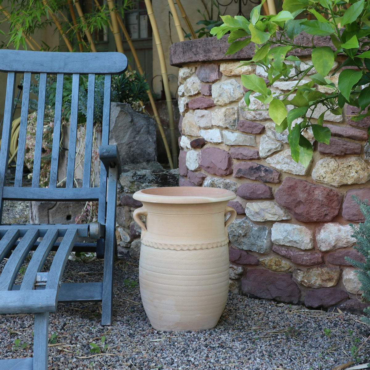 Eine ruhige Gartenszene zeigt einen blauen Holzstuhl neben der großen, leeren griechischen Amphorenvase „Cistus“, H=40cm. Der Sitzbereich ist von einer Steinmauer und üppigem grünem Laub umgeben und schafft so eine friedliche Außenumgebung. Der Boden ist mit Kies bedeckt und verleiht ihm eine rustikale Note.