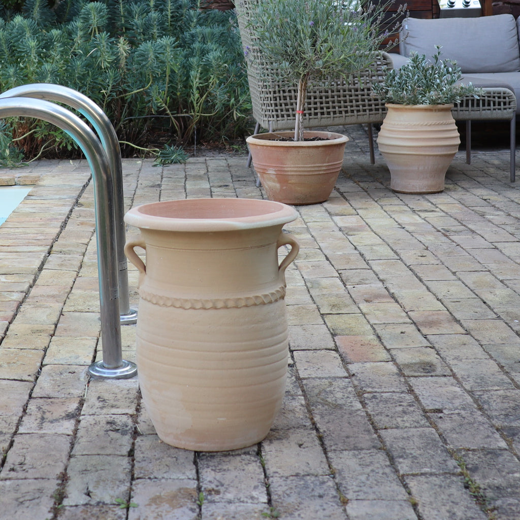 Auf einer Terrasse steht im Vordergrund eine große griechische Amphorenvase „Cistus“, H=40cm, mit Henkeln und im Hintergrund zwei weitere Töpfe mit Cistuspflanzen. Links davon ein Korbstuhl und ein silbernes Poolgeländer, umgeben von Grünpflanzen. Der Boden ist mit Steinfliesen gepflastert.
