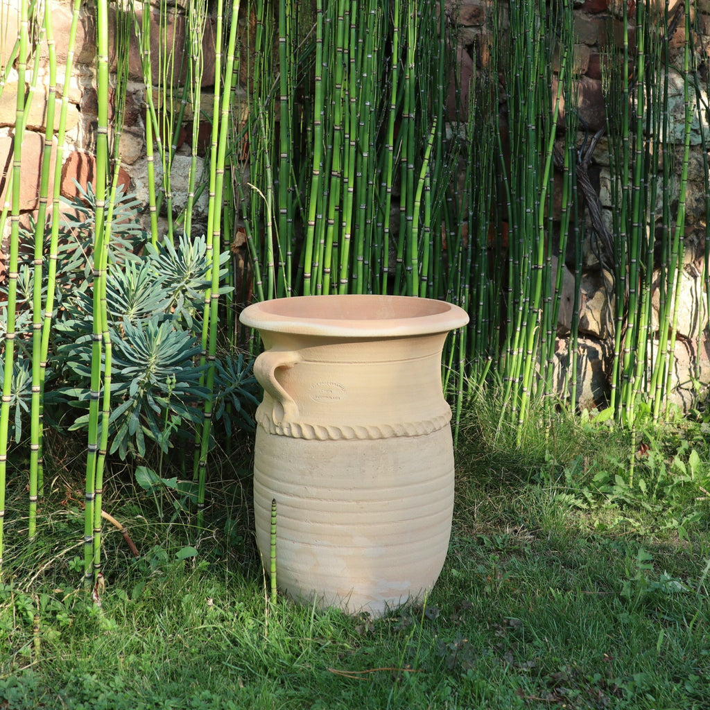 Eine große, beige griechische Amphorenvase aus Keramik „Cistus“, 40 cm hoch, mit dekorativen Griffen an beiden Seiten, steht auf einer Rasenfläche. Hohe, grüne Bambuspflanzen und eine Steinmauer dienen als Hintergrund und schaffen eine natürliche und ruhige Umgebung. Sonnenlicht wirft weiche Schatten, die die ruhige Atmosphäre verstärken, während in der Nähe Zistrosen blühen.